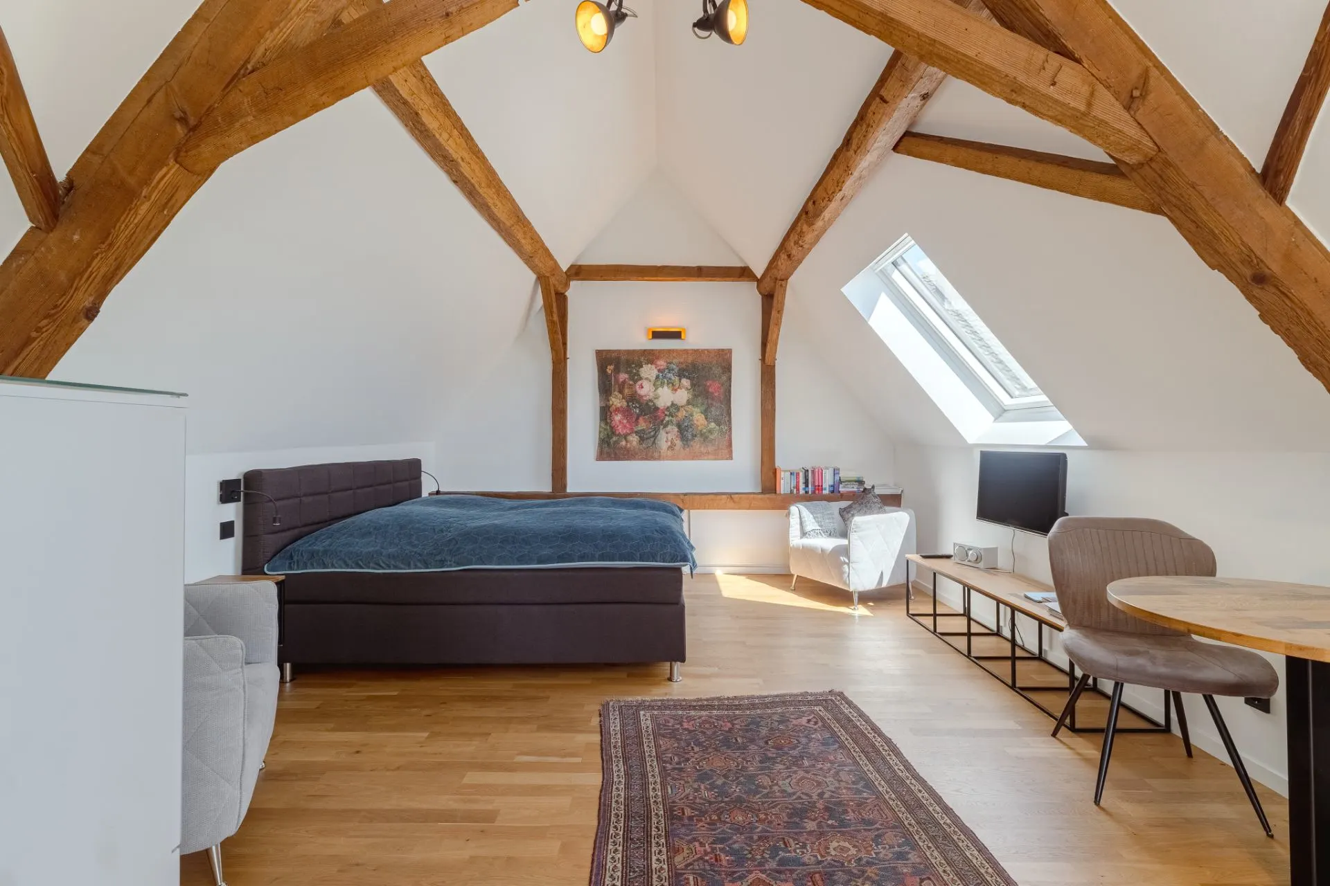 Helles Dachgeschosszimmer mit Holzbalken, Bett, Teppich, Sitzecke und Fenster.