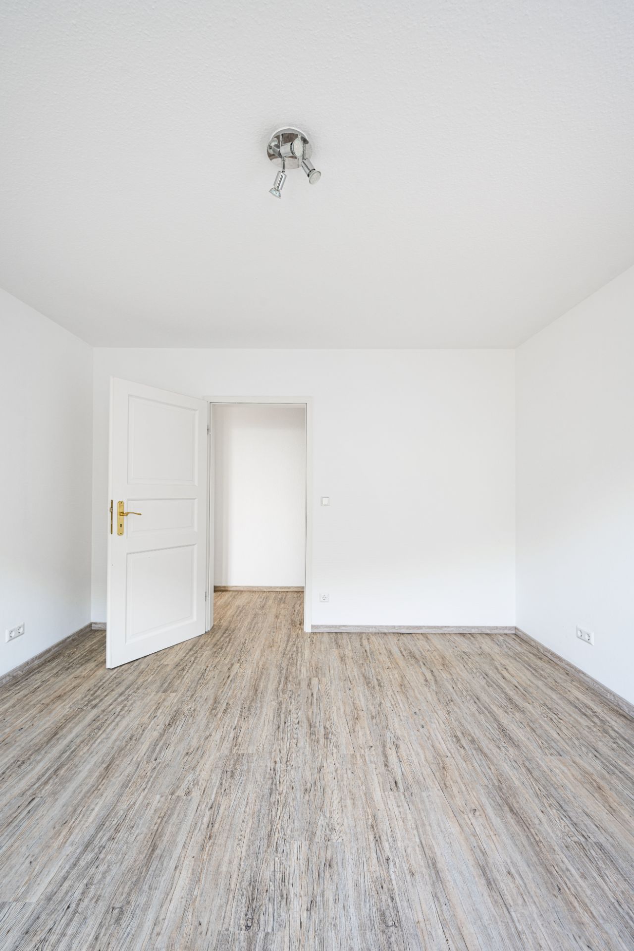 Leerer Raum mit Holzfußboden, weißen Wänden, Deckenleuchte und geöffneter Tür.