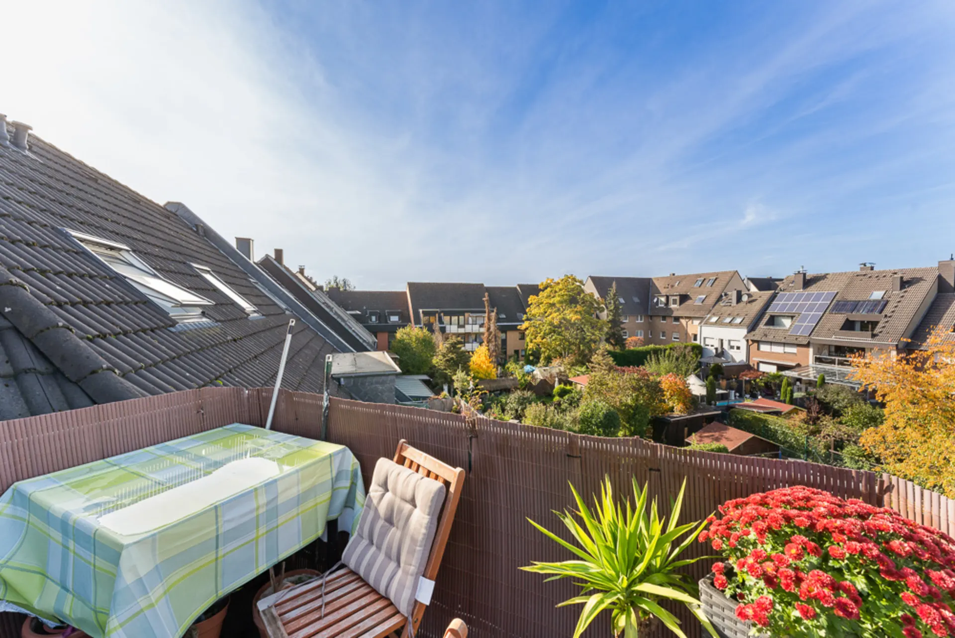 Balkon mit Tisch, zwei Stühlen und Blumen, Blick auf Häuser und Gärten bei sonnigem Wetter.
