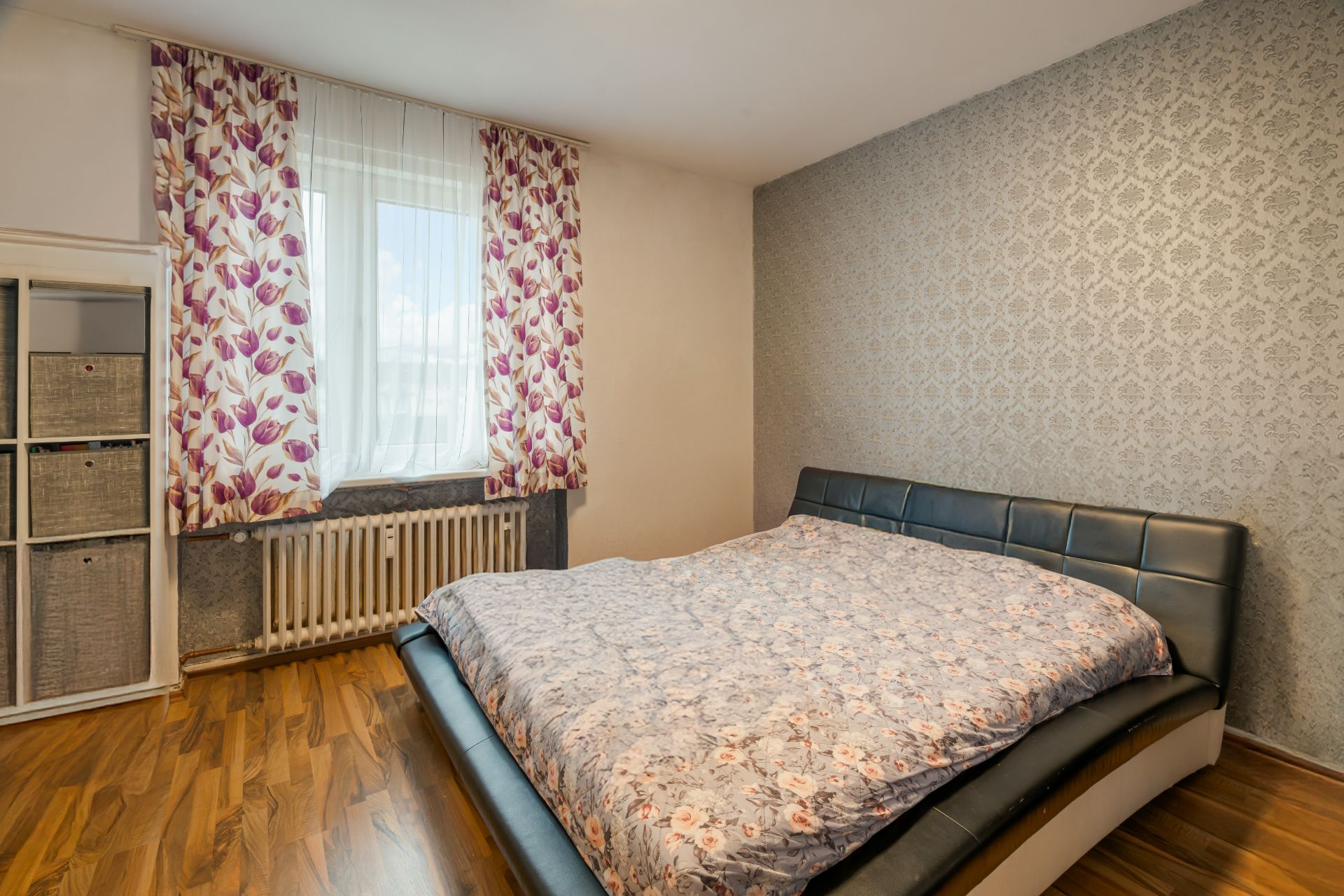 Schlafzimmer mit Doppelbett, floralem Vorhang und Regal mit Körben auf Holzfußboden.