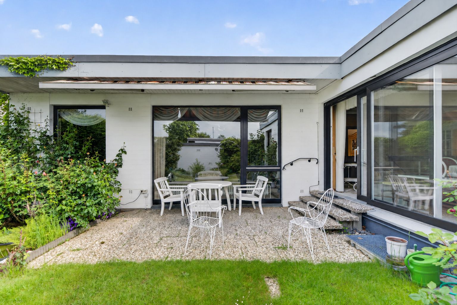 Terrasse mit weißen Gartenmöbeln, großen Fenstern und angrenzendem Gartenbereich.