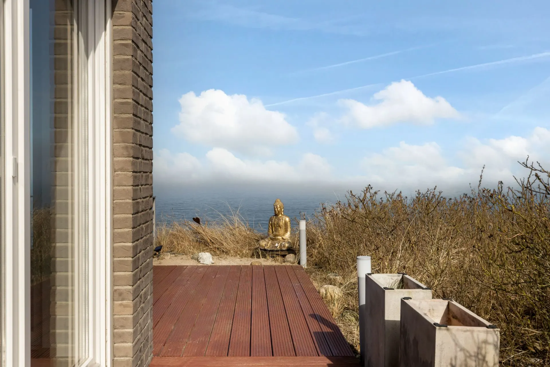 Holzterrasse mit Blick auf das Meer, goldene Buddhastatue und trockene Sträucher im Hintergrund.