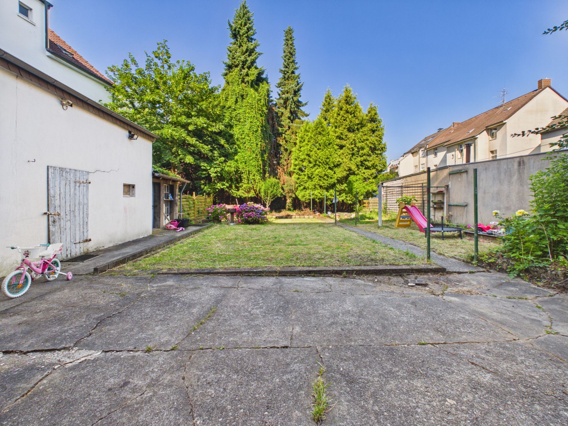 Hof mit Betonfläche, Rasen, Bäumen, Rutsche und Kinderfahrrad neben einem Wohnhaus.