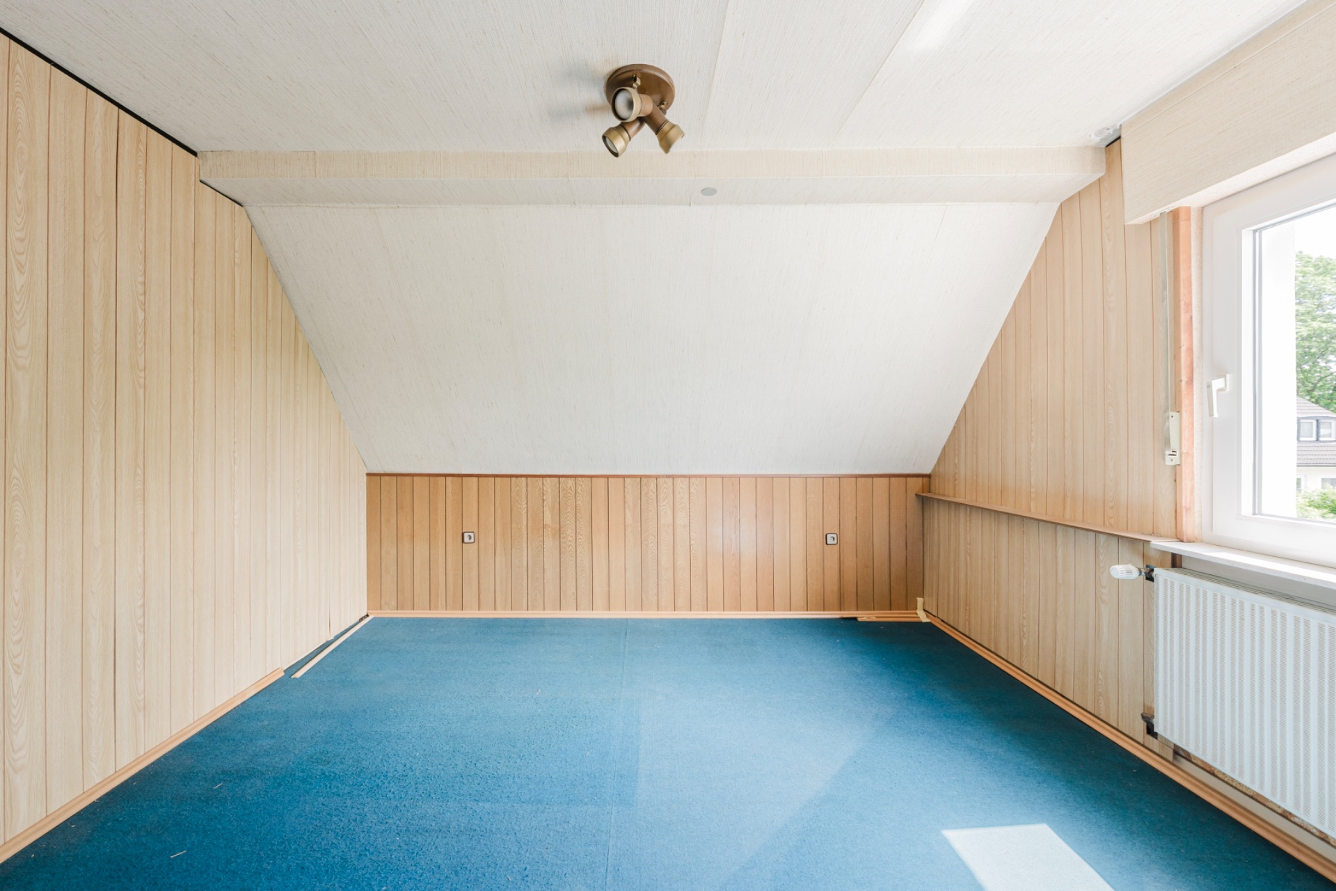 Leeres Zimmer mit blauer Teppichboden, Holzverkleidung und Fenster rechts.