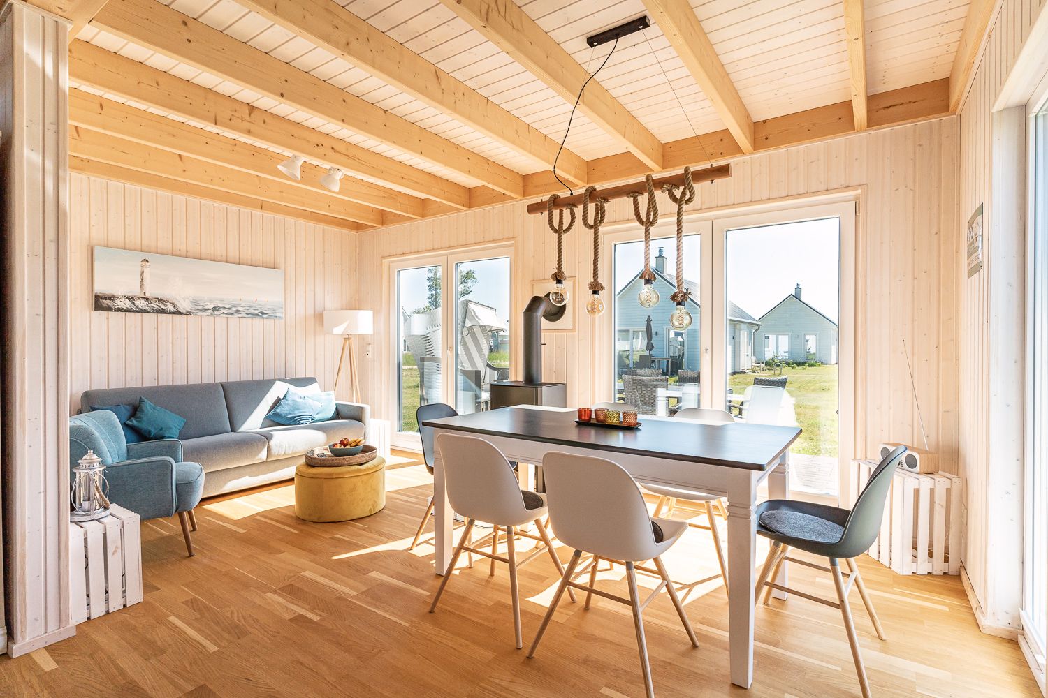 Helles Wohnzimmer mit Holzboden, Esstisch, Stühlen und Sofa, große Fenster mit Blick nach draußen.