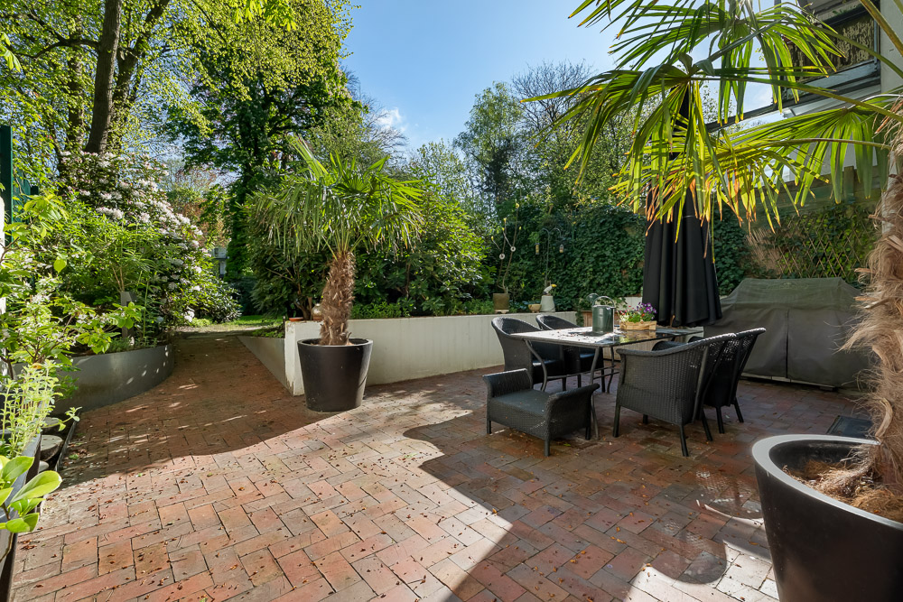 Gepflasterte Terrasse mit Gartenmöbeln, Palmen in Töpfen und viel Grün im Hintergrund.