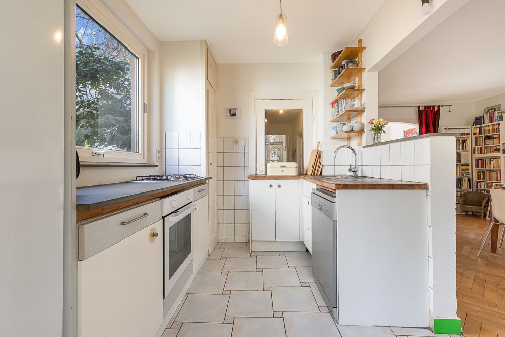 Helle Einbauküche mit Holzarbeitsplatten, Fenster, offenem Regal und Blick ins Wohnzimmer.