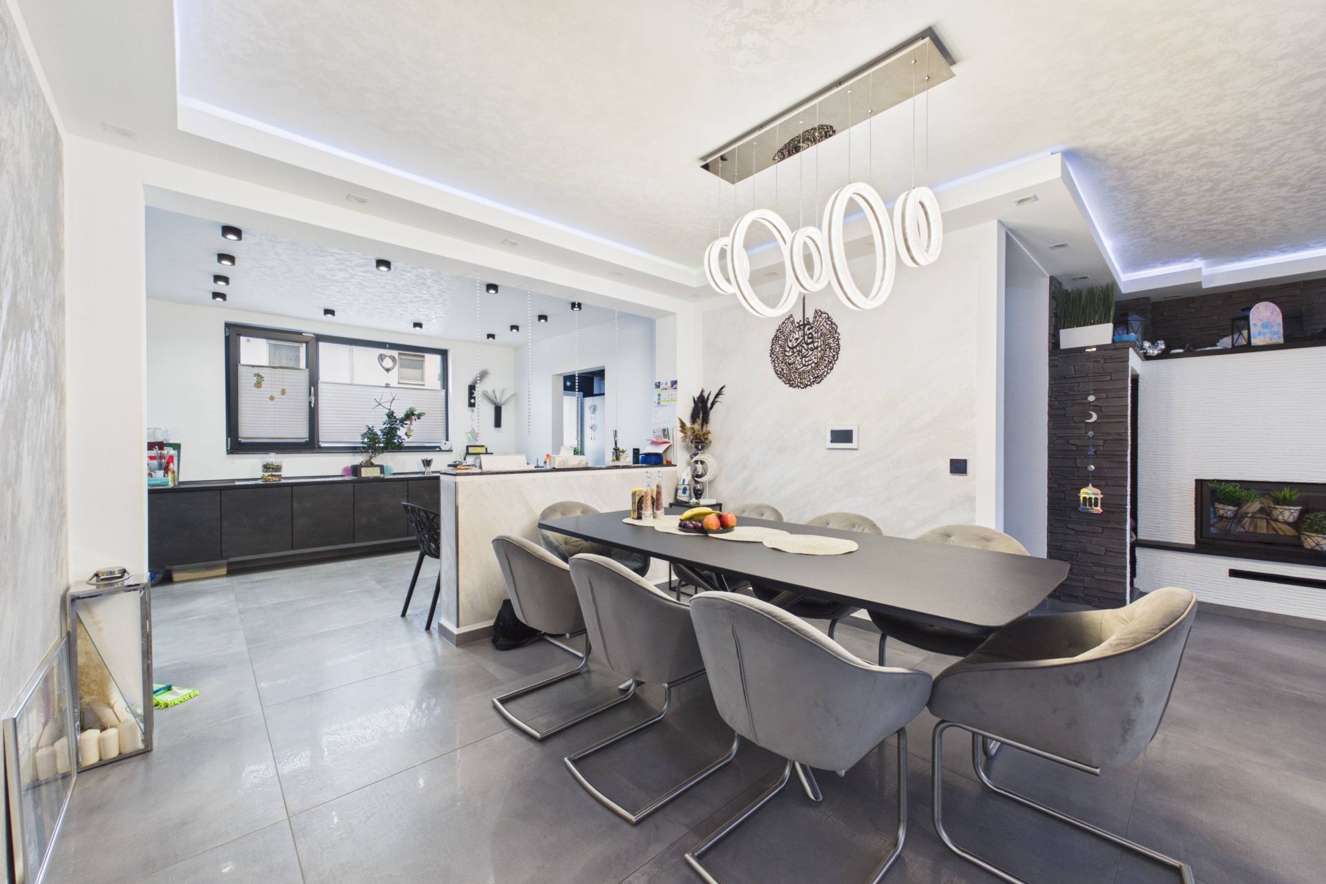 Modernes Esszimmer mit großem Tisch, grauen Stühlen und auffälliger Lampe, offene Küche im Hintergrund.