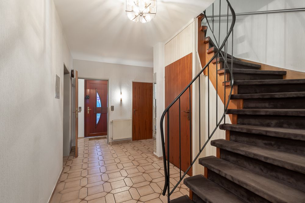 Flur mit brauner Haustür, gefliestem Boden, Treppe mit Teppich und mehreren Holztüren.
