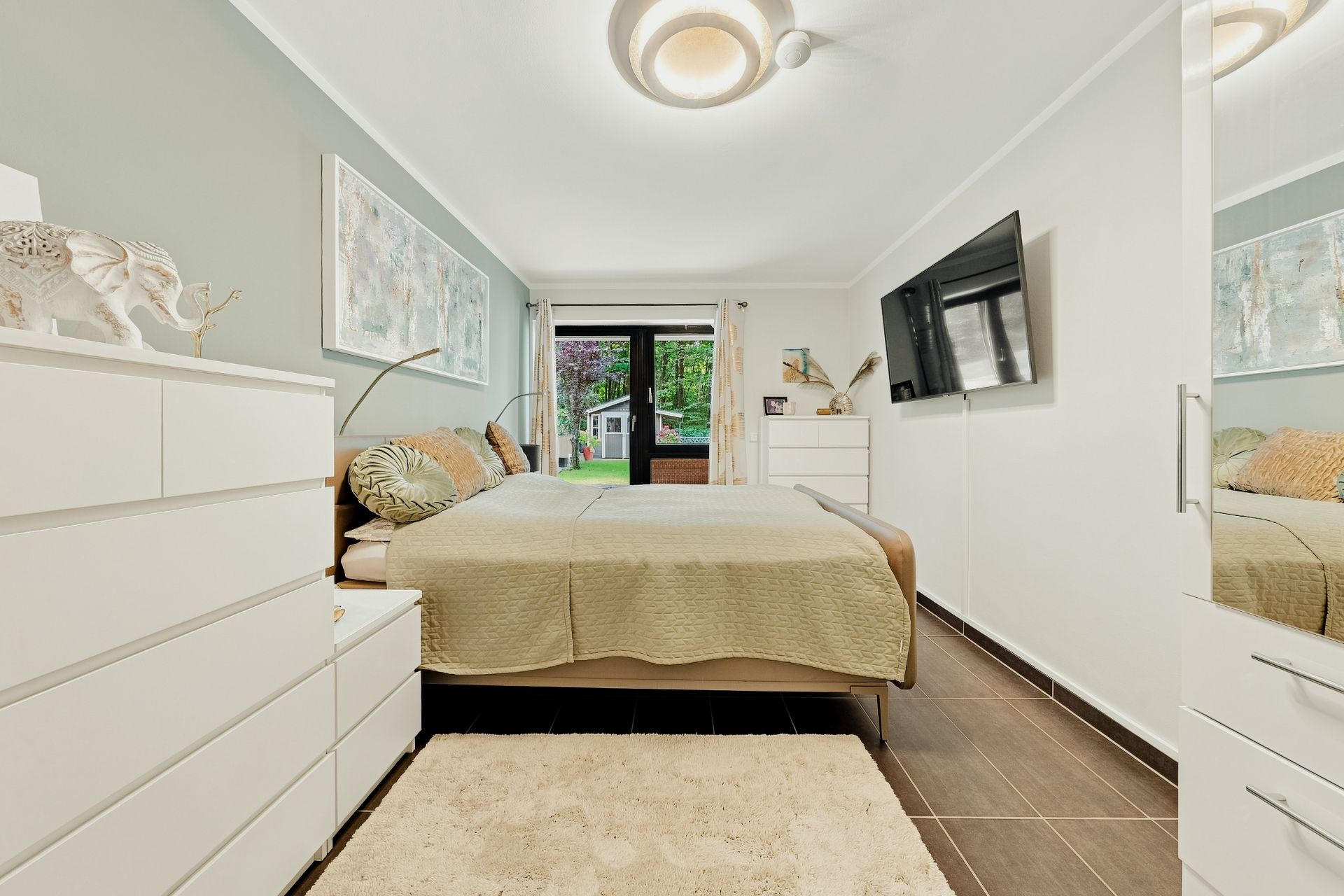 Modernes Schlafzimmer mit Doppelbett, Kommode, Fernseher an der Wand und Blick auf den Garten.