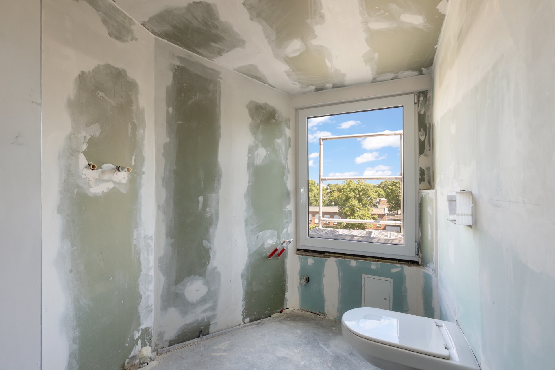 Unfertiges Badezimmer mit WC und großem Fenster, nackten Wänden und sichtbaren Anschlüssen.