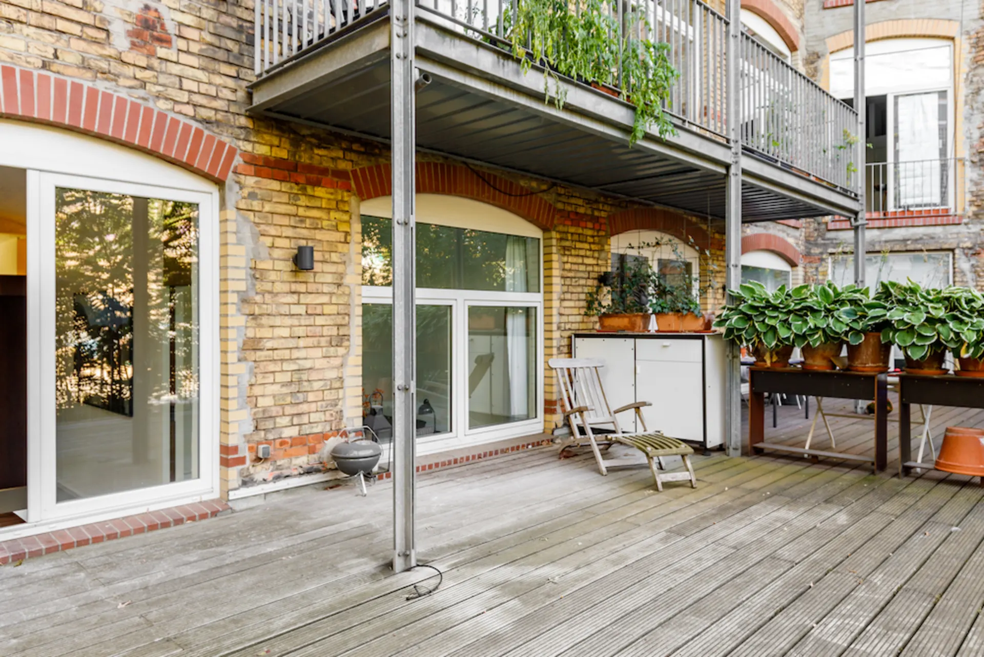 Große Holzterrasse mit Pflanzen, Stühlen und Zugang zu einer Wohnung in einem Backsteingebäude.