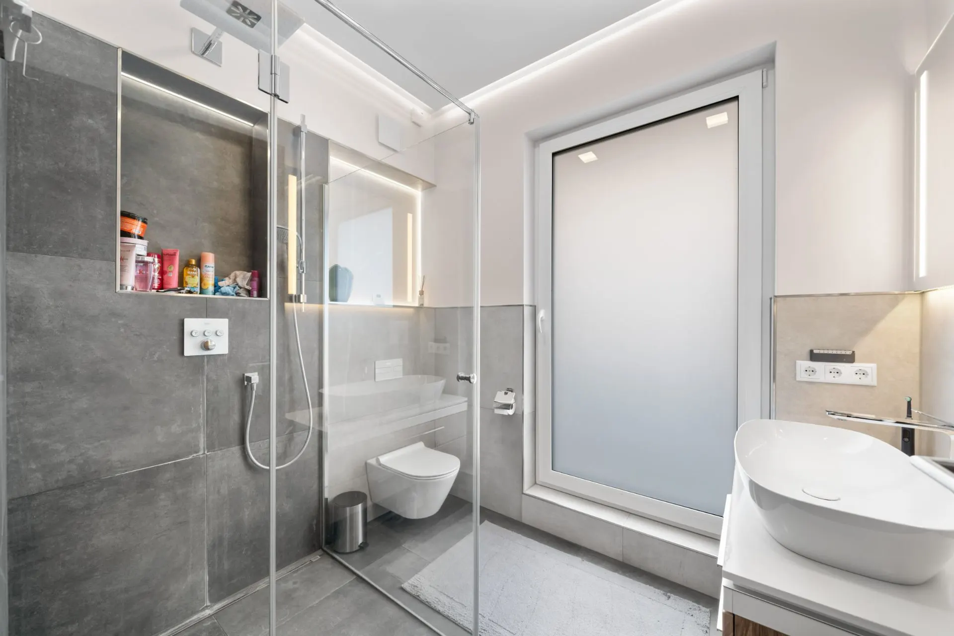 Modernes Badezimmer mit großer Dusche, WC, Fenster und Waschbecken mit Spiegel.