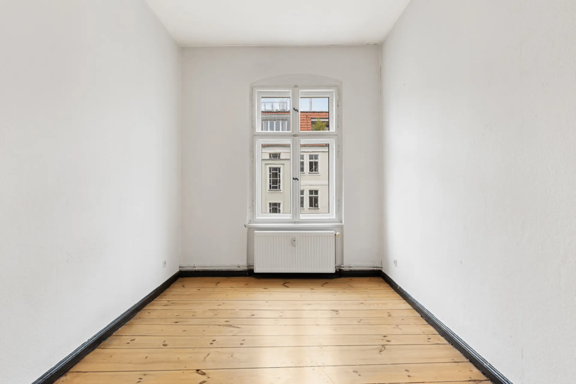 Heller, leerer Raum mit Holzboden, weiß gestrichenen Wänden und einem Fenster mit Heizkörper darunter.