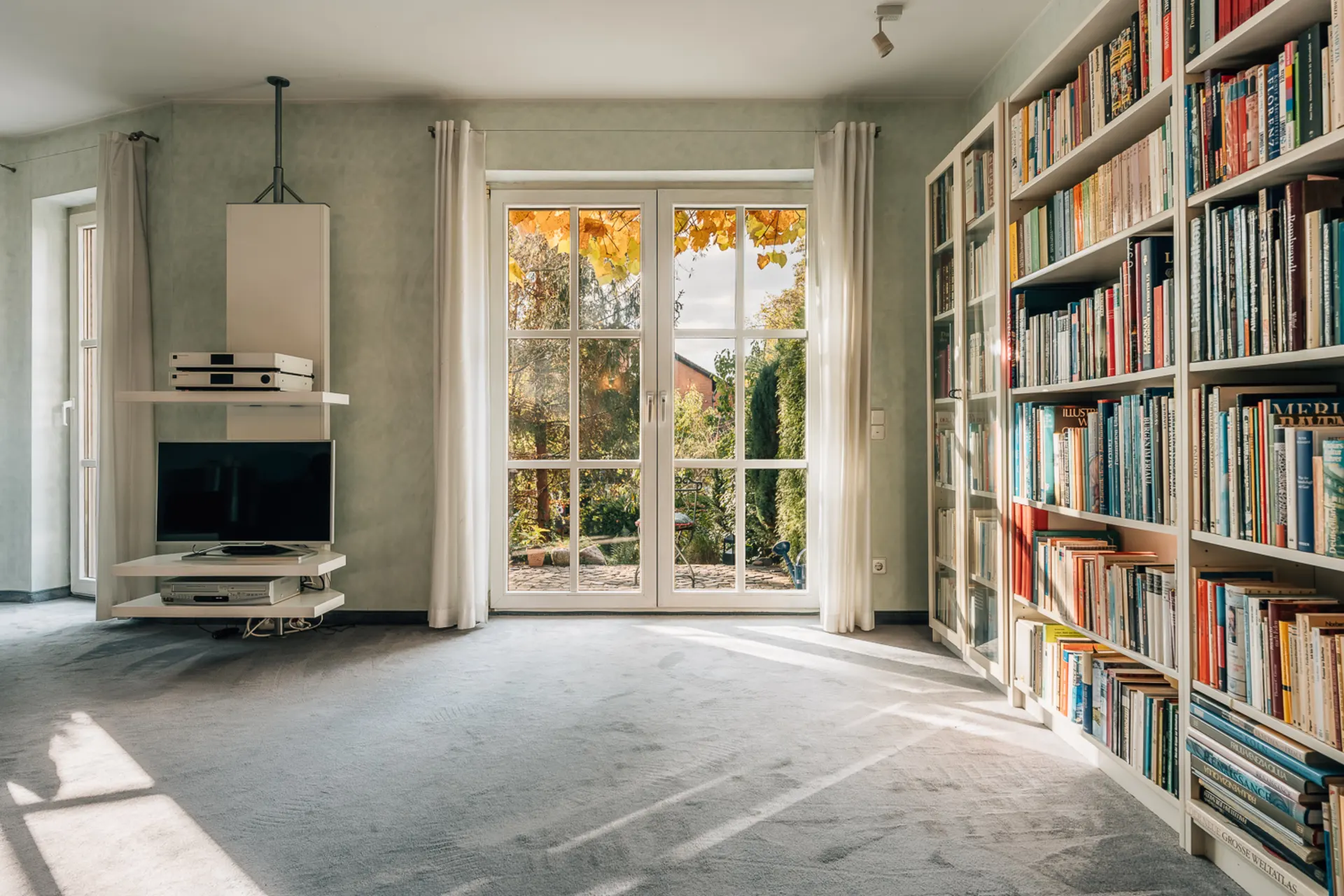 Helles Wohnzimmer mit Teppich, Bücherregal, Glastüren zum Garten und kleinem Fernseher.