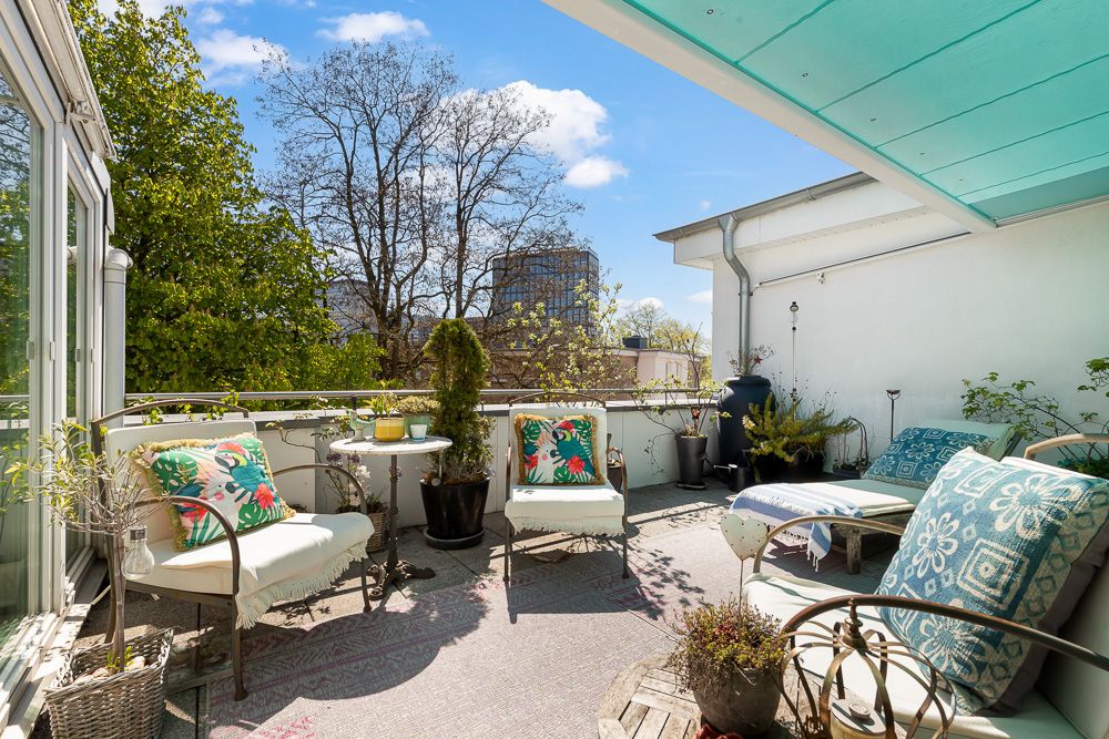 Sonnige Terrasse mit Sitzgelegenheiten, Pflanzen und kleinem Tisch, umgeben von Bäumen.