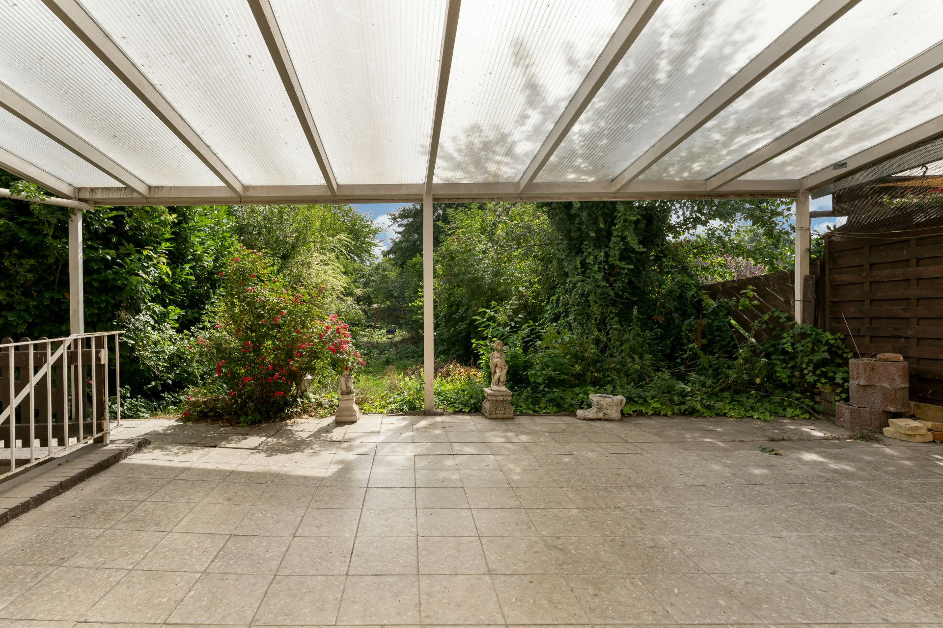 Überdachte Terrasse mit Fliesenboden, Blick auf Garten mit Pflanzen und Skulpturen.