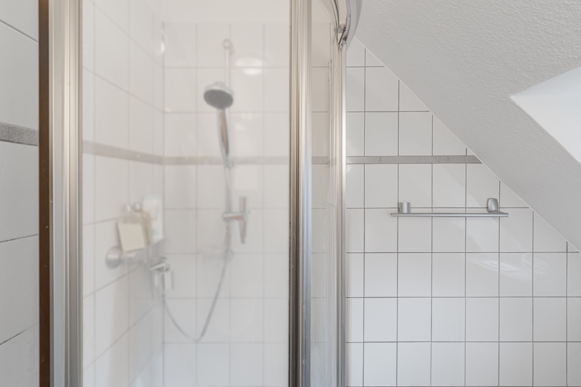 Moderne, weiß geflieste Dusche mit Glasabtrennung in einem Bad unter einer Dachschräge.