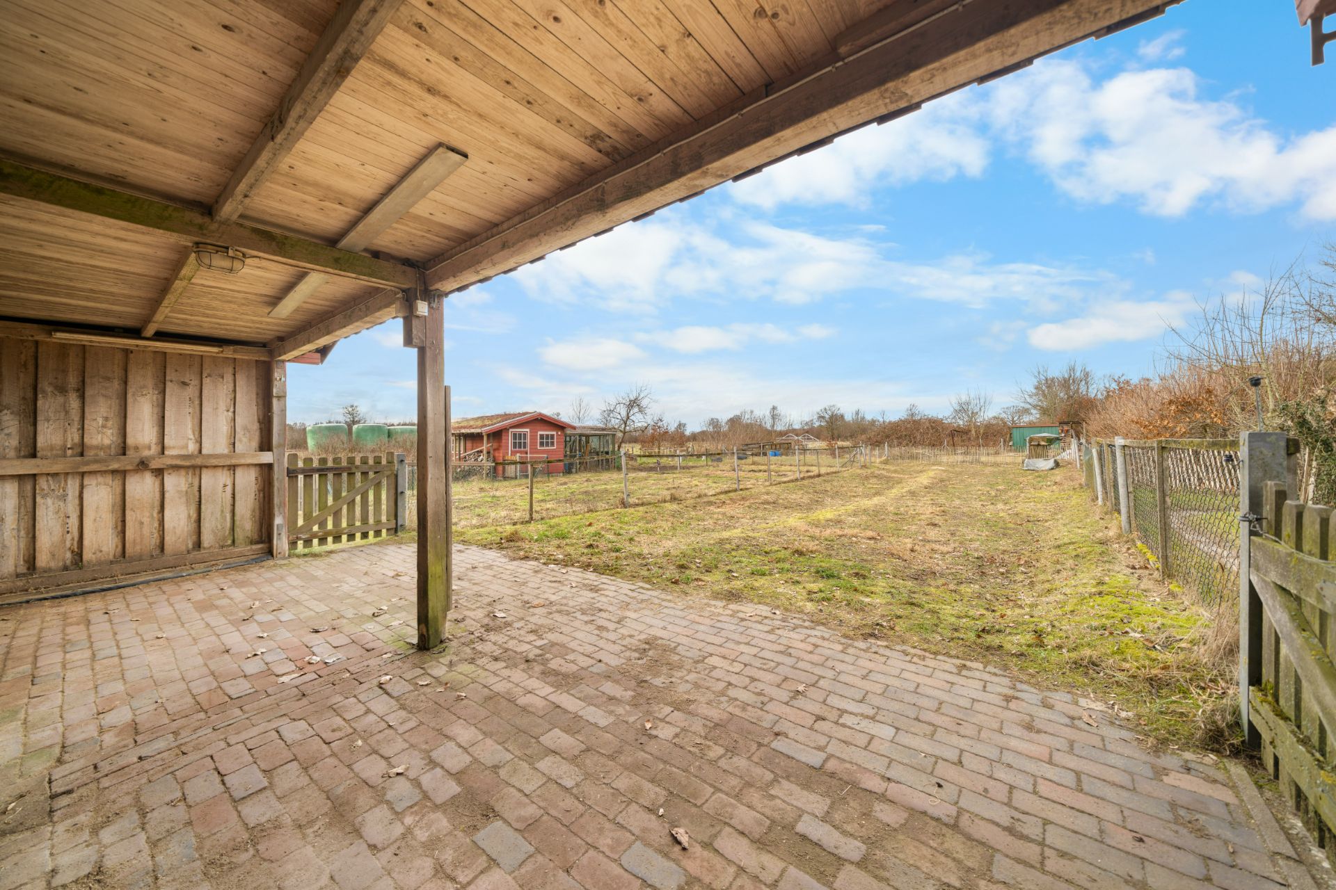 Überdachte Terrasse mit Blick auf einen Garten und ein rotes Gartenhaus im Hintergrund.