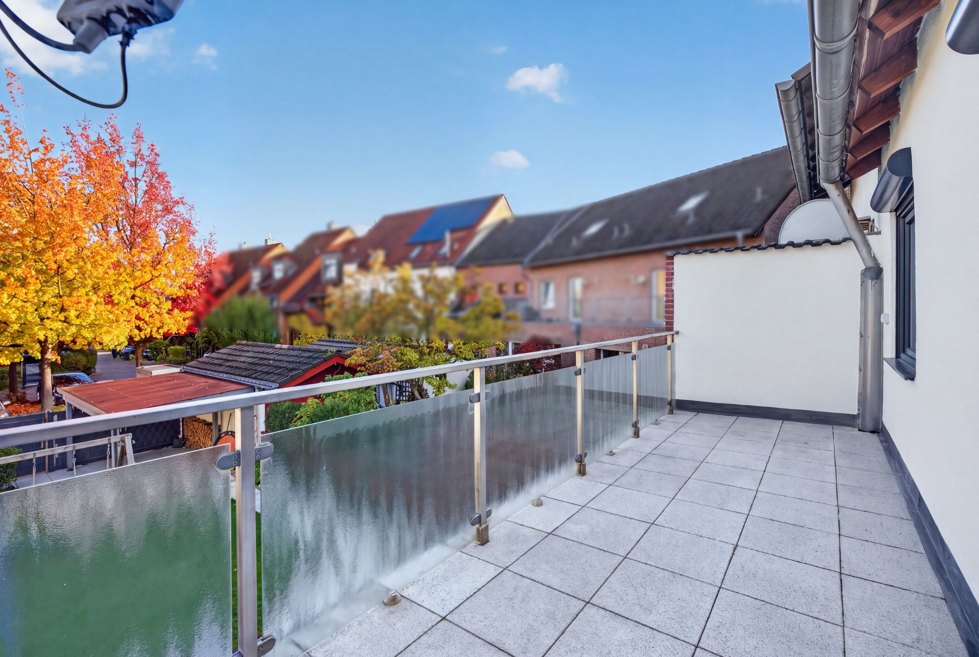 Großer Balkon mit Glasgeländer und Blick auf Nachbarhäuser und Bäume im Herbst.