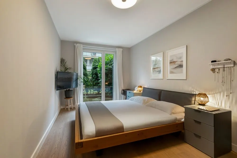 Modernes Schlafzimmer mit Doppelbett, Fernseher, Nachttischen und Blick auf einen Garten.