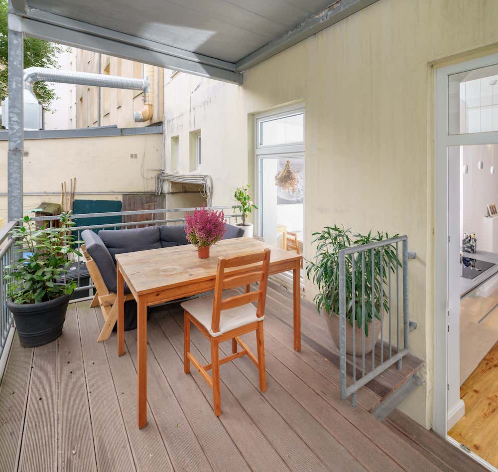 Überdachter Balkon mit Holztisch, Stuhl, grauem Sofa und mehreren Pflanzen in Töpfen.