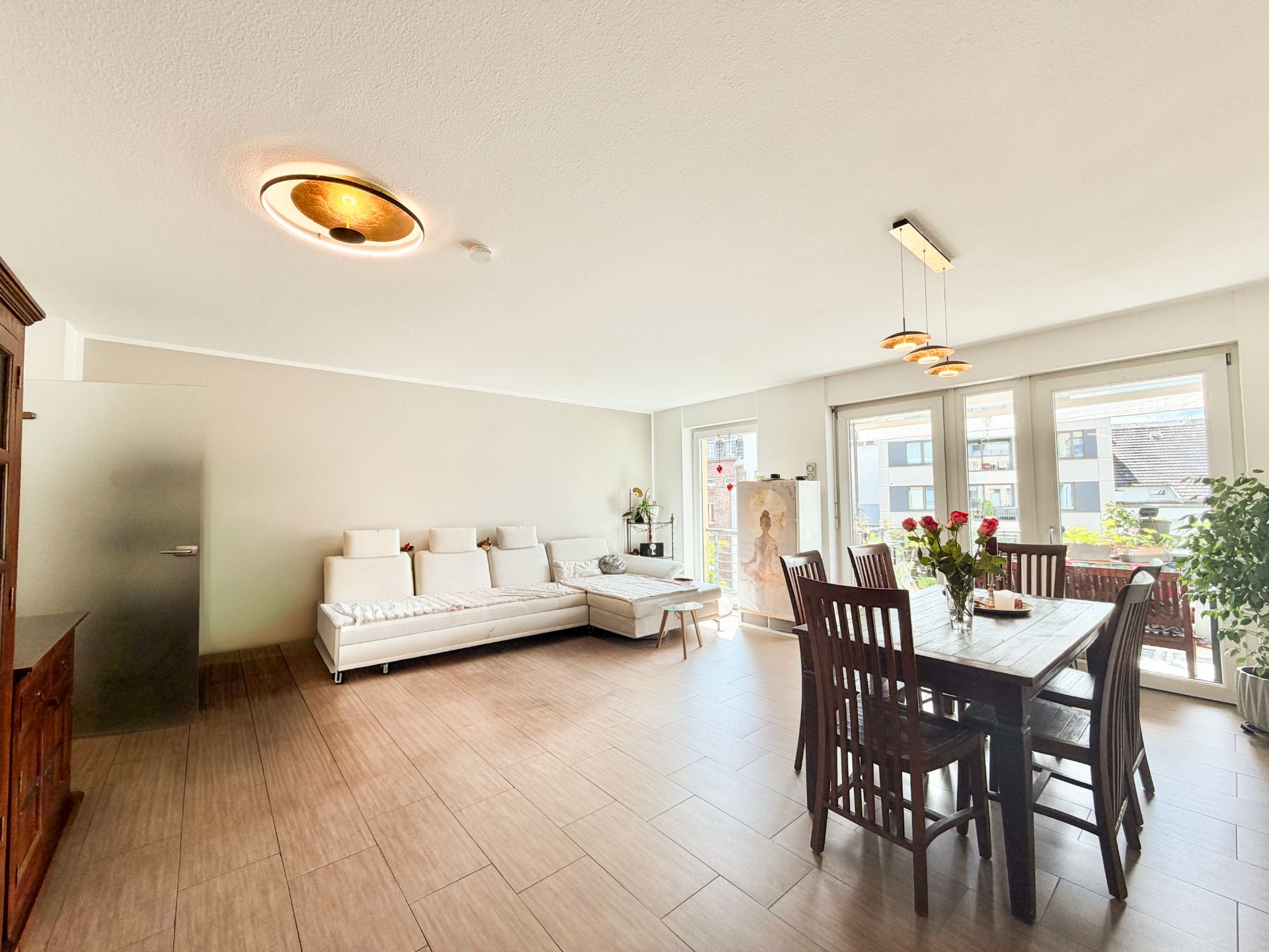Helles Wohnzimmer mit Ecksofa, Esstisch mit Stühlen und großen Fenstern zu einem Balkon.