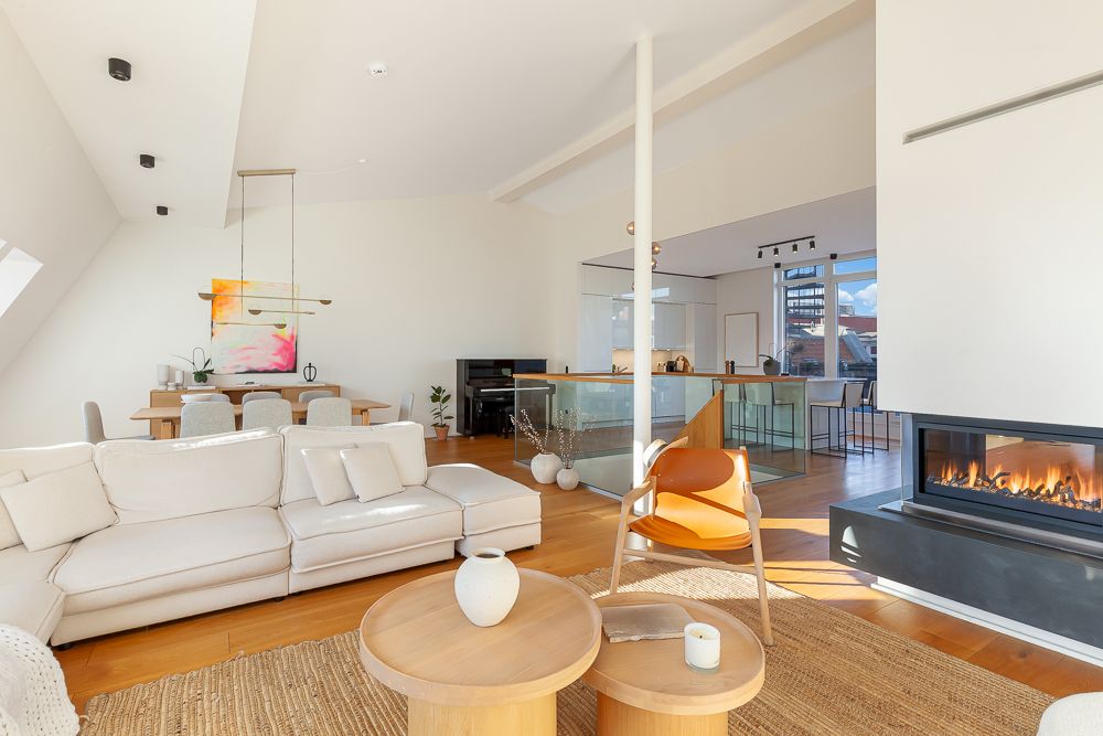 Helles Wohnzimmer mit weißem Sofa, Kamin, Holztisch und moderner offener Küche im Hintergrund.