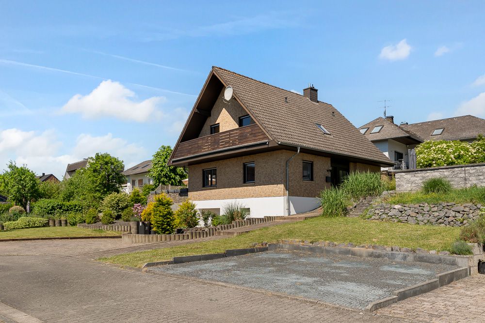 Einfamilienhaus mit Satteldach, Garten und gepflastertem Parkplatz davor bei sonnigem Wetter.