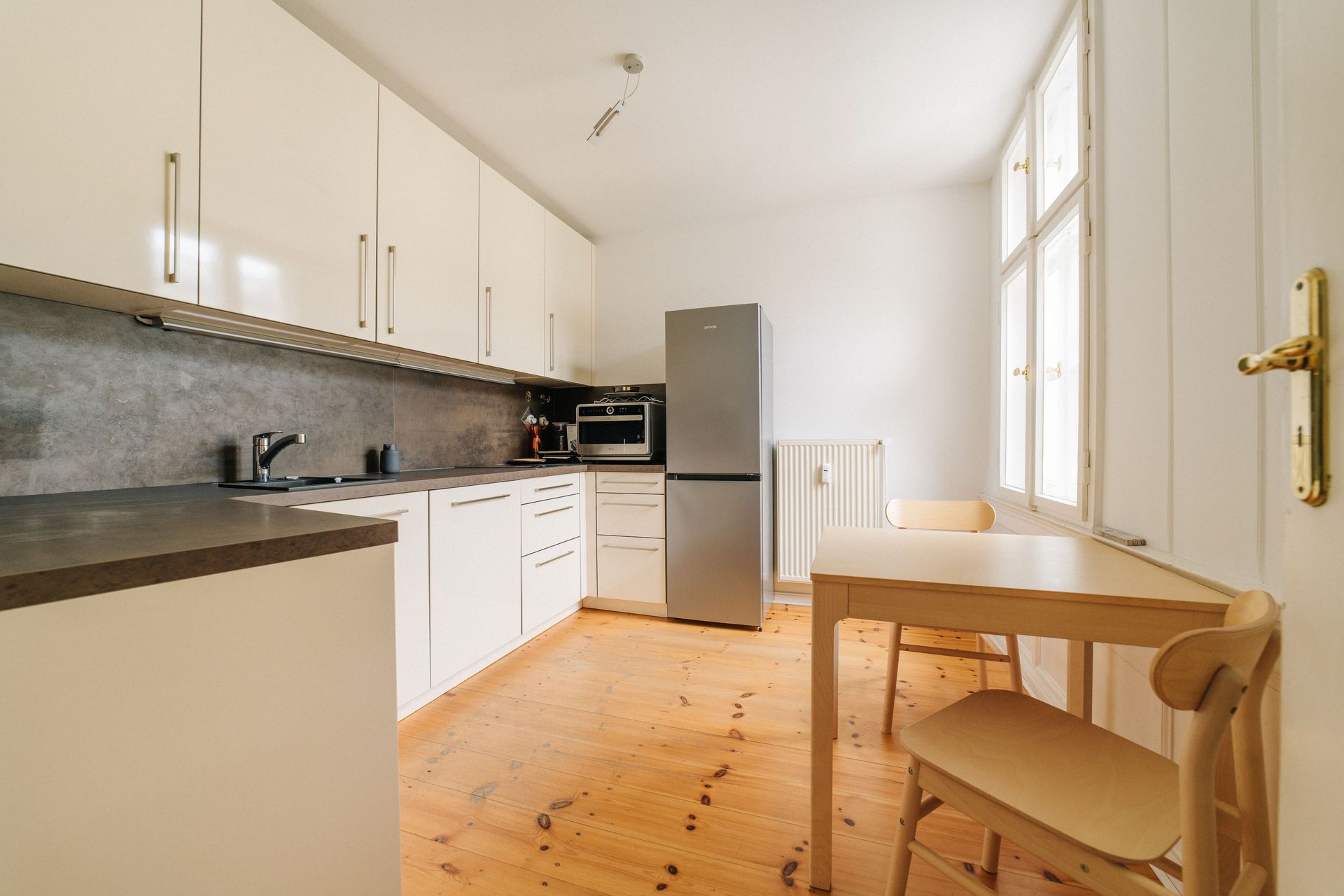 Helle Küche mit weißen Schränken, Holzfußboden, kleinem Esstisch und großem Fenster.