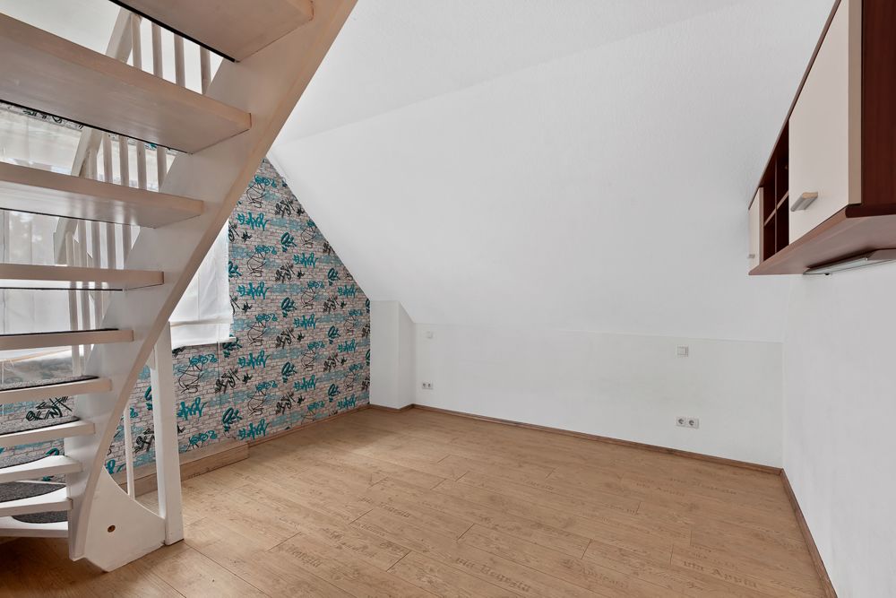 Leeres Zimmer mit Holzfußboden, weißer Treppe und gemusterter Tapete an einer Wand.
