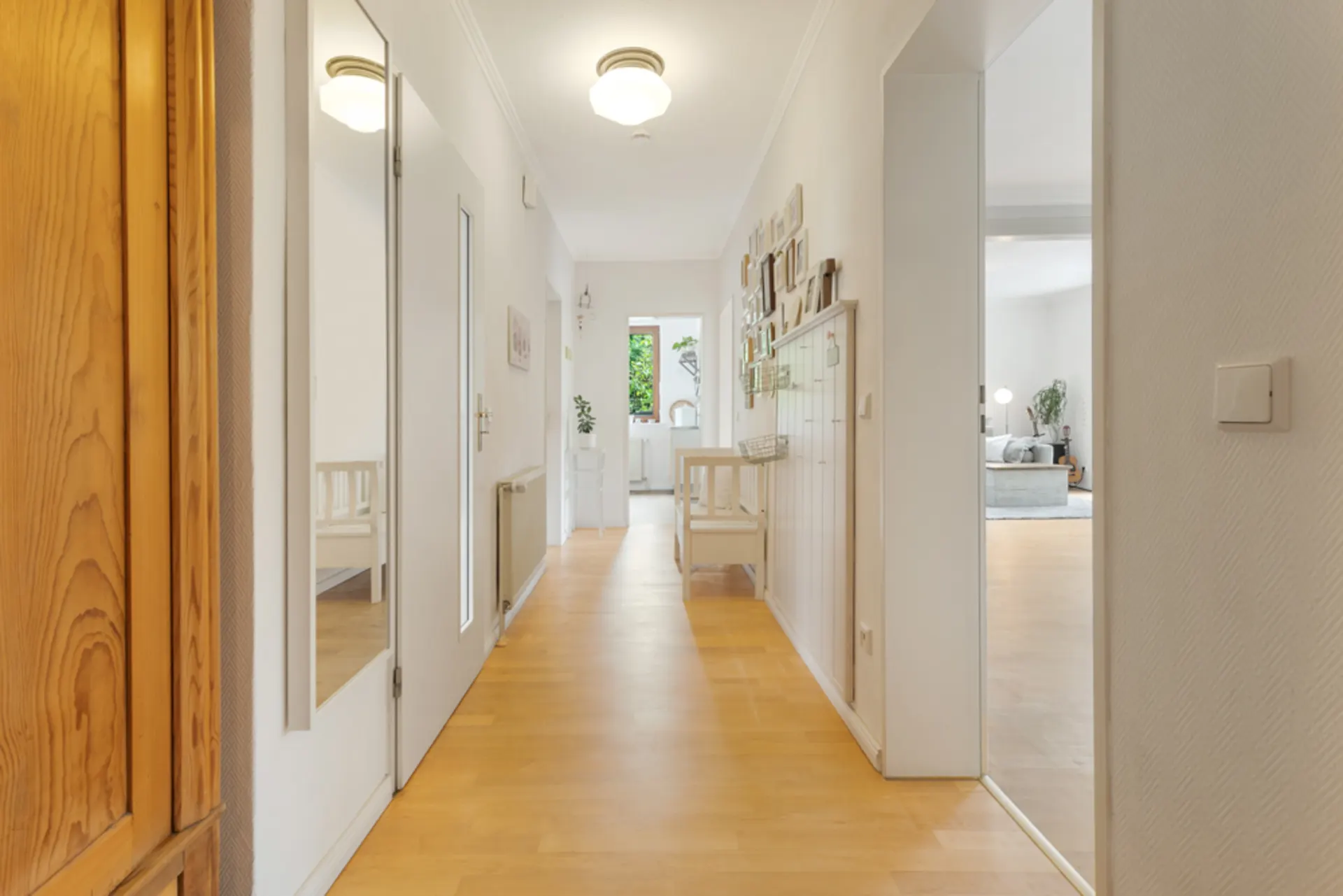 Heller Flur mit Holzfußboden, weißen Wänden und Spiegel, Blick in einen Wohnraum und Küche.