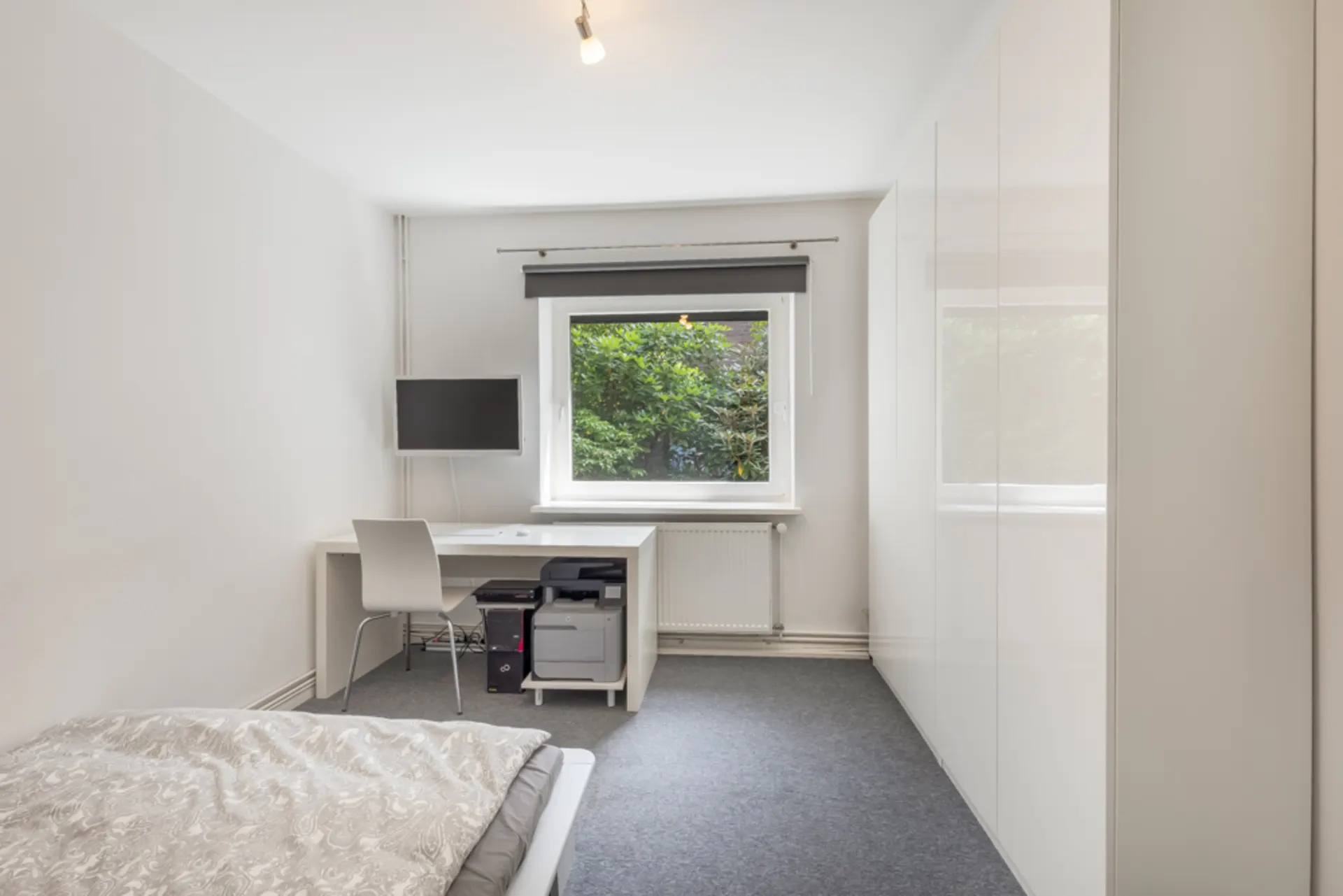 Helles Schlafzimmer mit Bett, Schreibtisch, Stuhl und großem Fenster mit Blick ins Grüne.