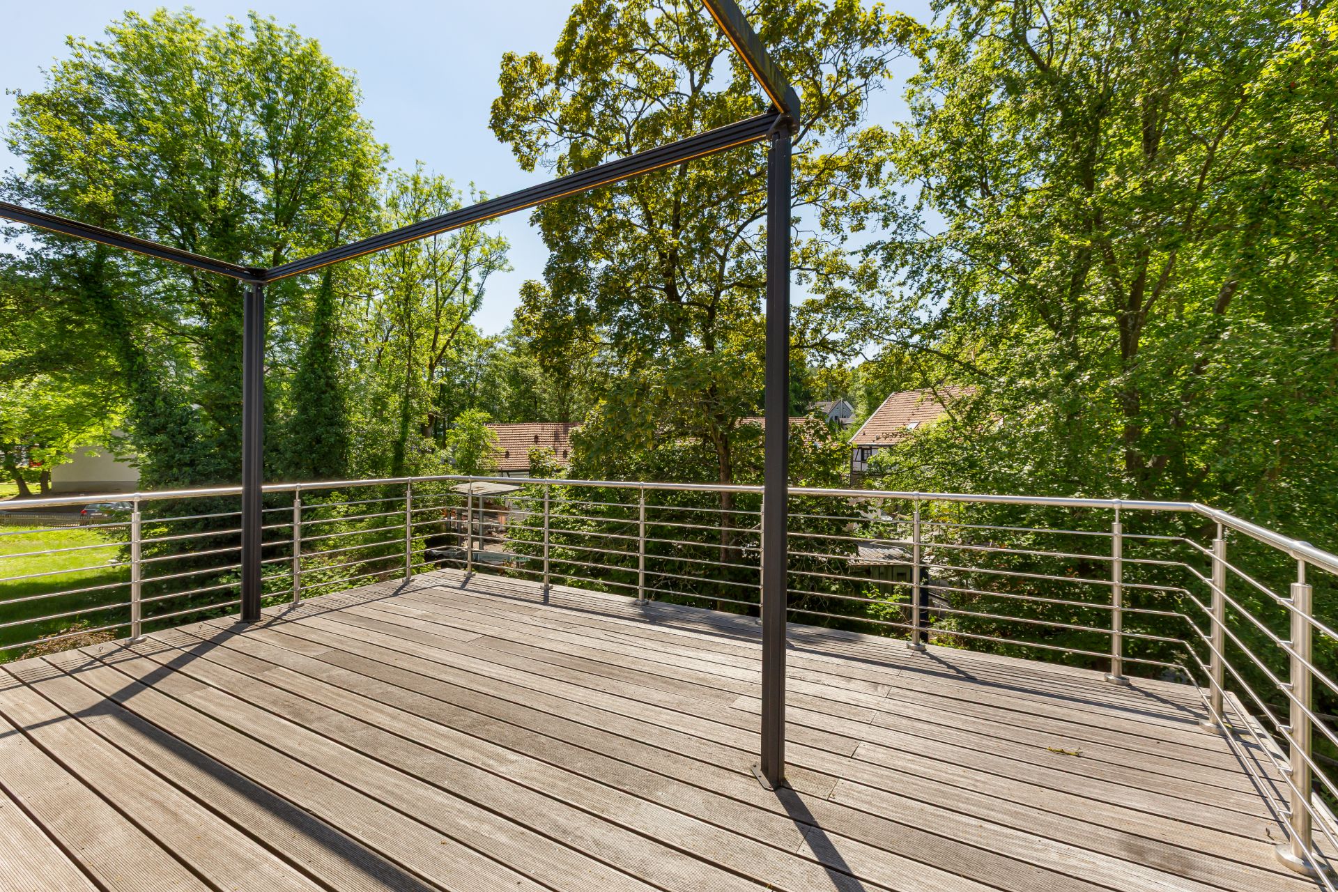 Holzterrasse mit Metallgeländer und Rahmen, umgeben von grünen Bäumen.