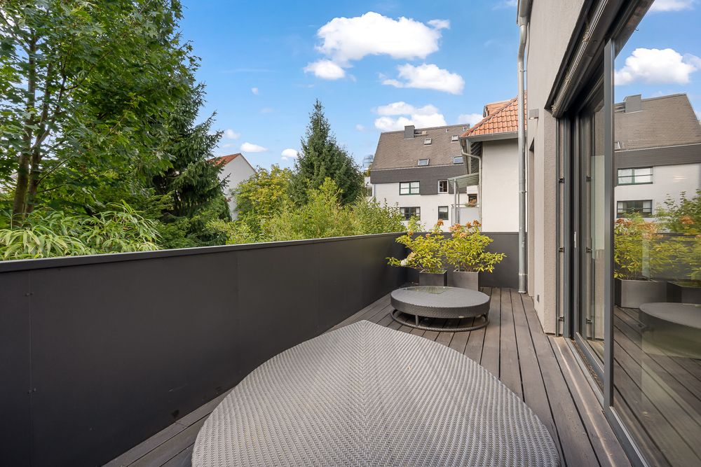 Moderne Holz-Terrasse mit runden Gartenmöbeln und Pflanzen, umgeben von Bäumen und Nachbarhäusern.