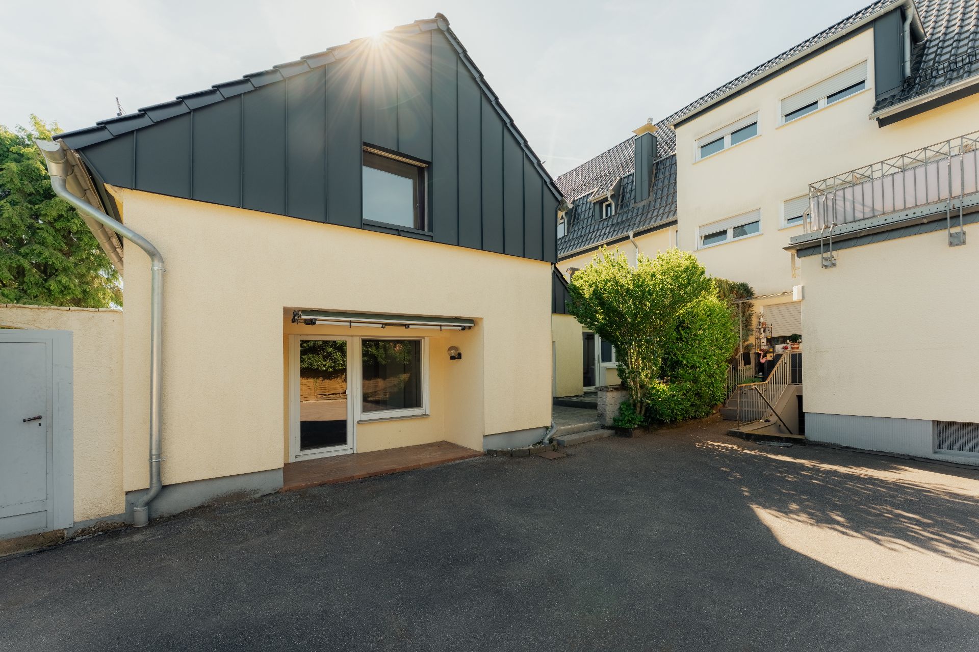 Zweistöckiges Wohnhaus mit gelber Fassade, großem Fenster und Asphaltfläche im Vordergrund.