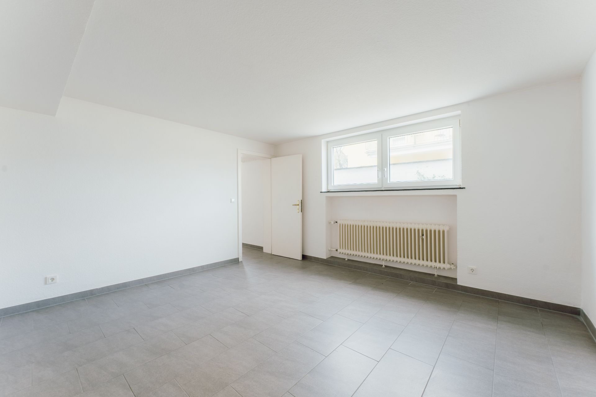Heller leerer Raum mit grauem Fliesenboden, weißer Wand, Fenster und Heizkörper.