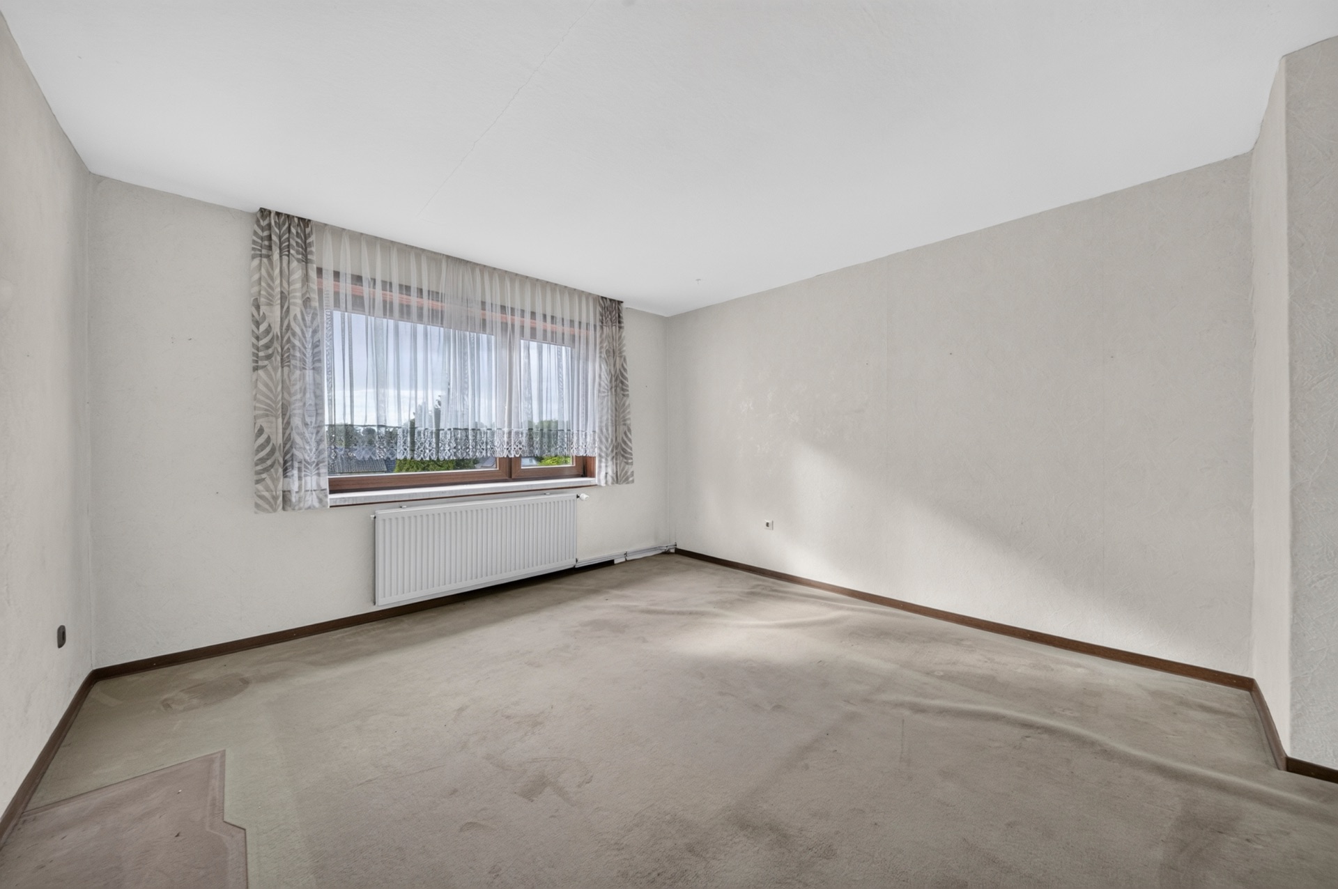 Leerer Raum mit Teppichboden, großem Fenster mit Vorhang und Heizkörper unter dem Fenster.