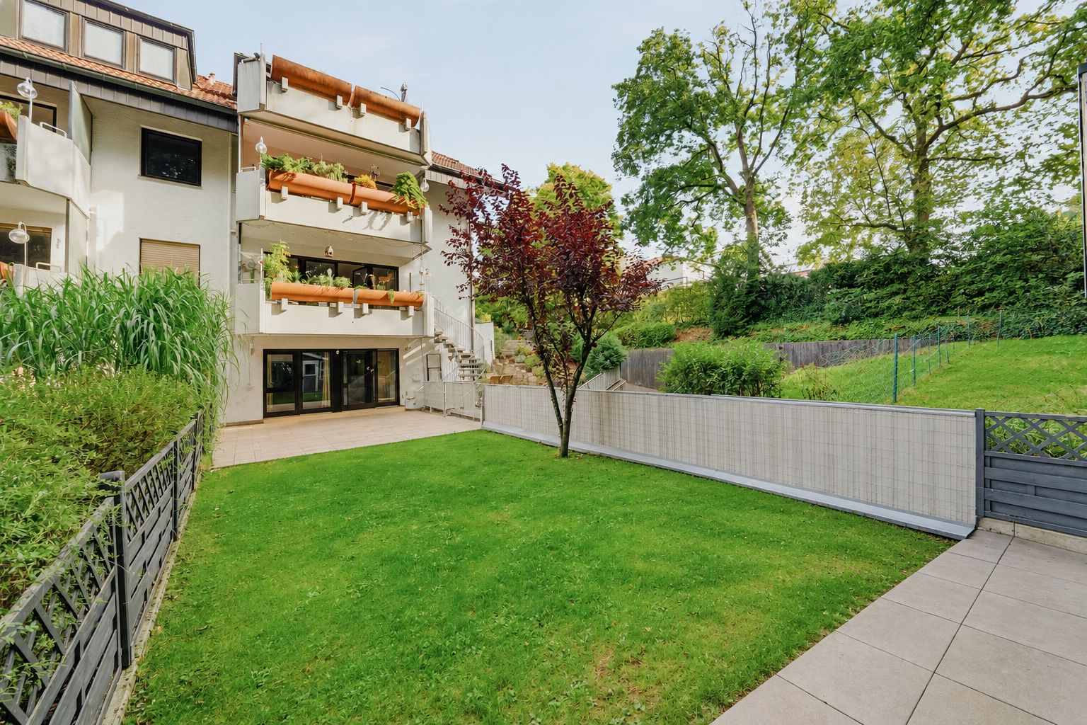 Grüner Garten mit Baum, Zaun und moderner Wohnhausfassade mit Balkonen im Hintergrund.