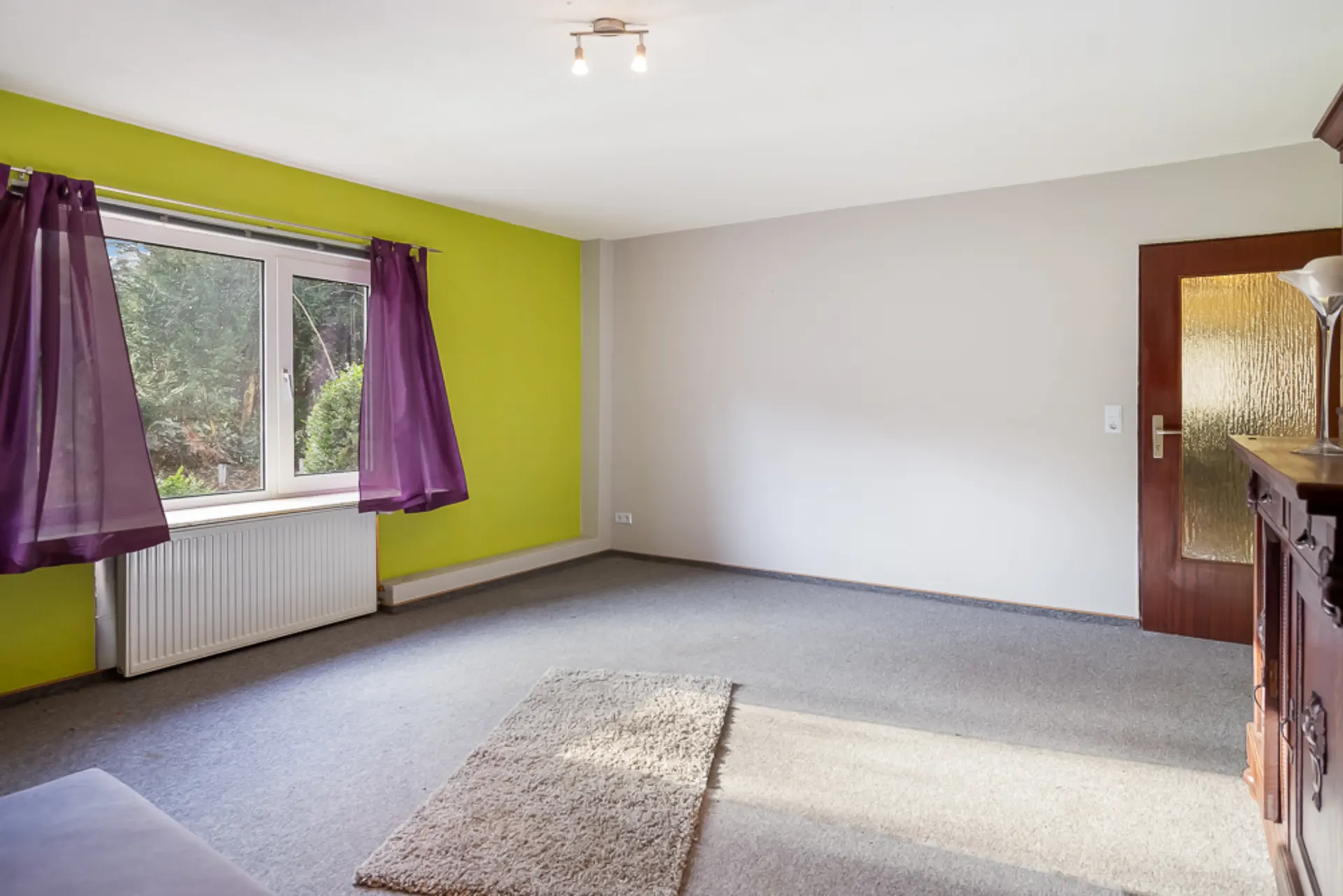 Helles Wohnzimmer mit grün gestrichener Wand, großen Fenstern und lila Vorhängen.