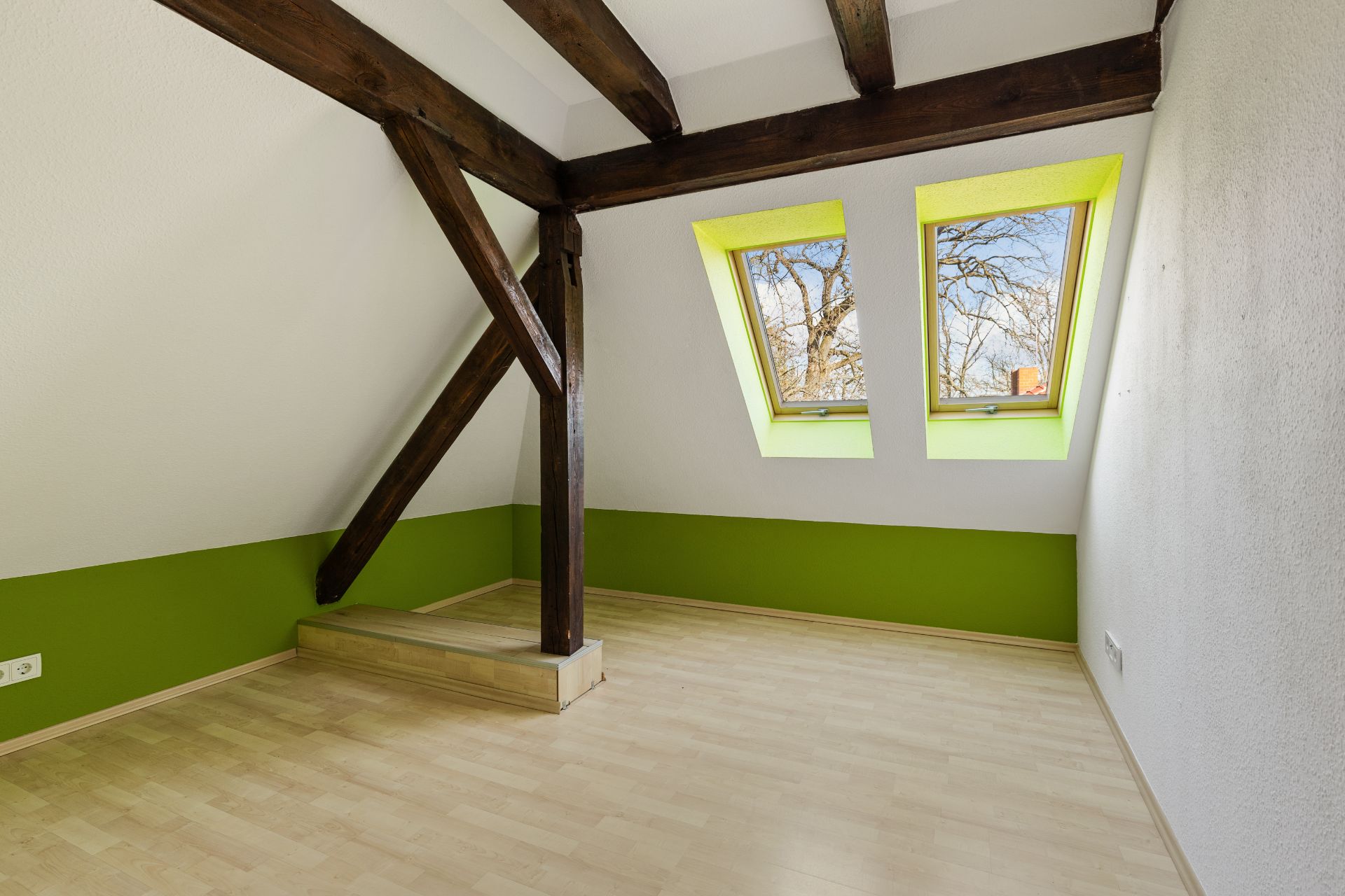Leerer Dachzimmer mit Holzträgern, grünem Wandstreifen und zwei Dachfenstern.