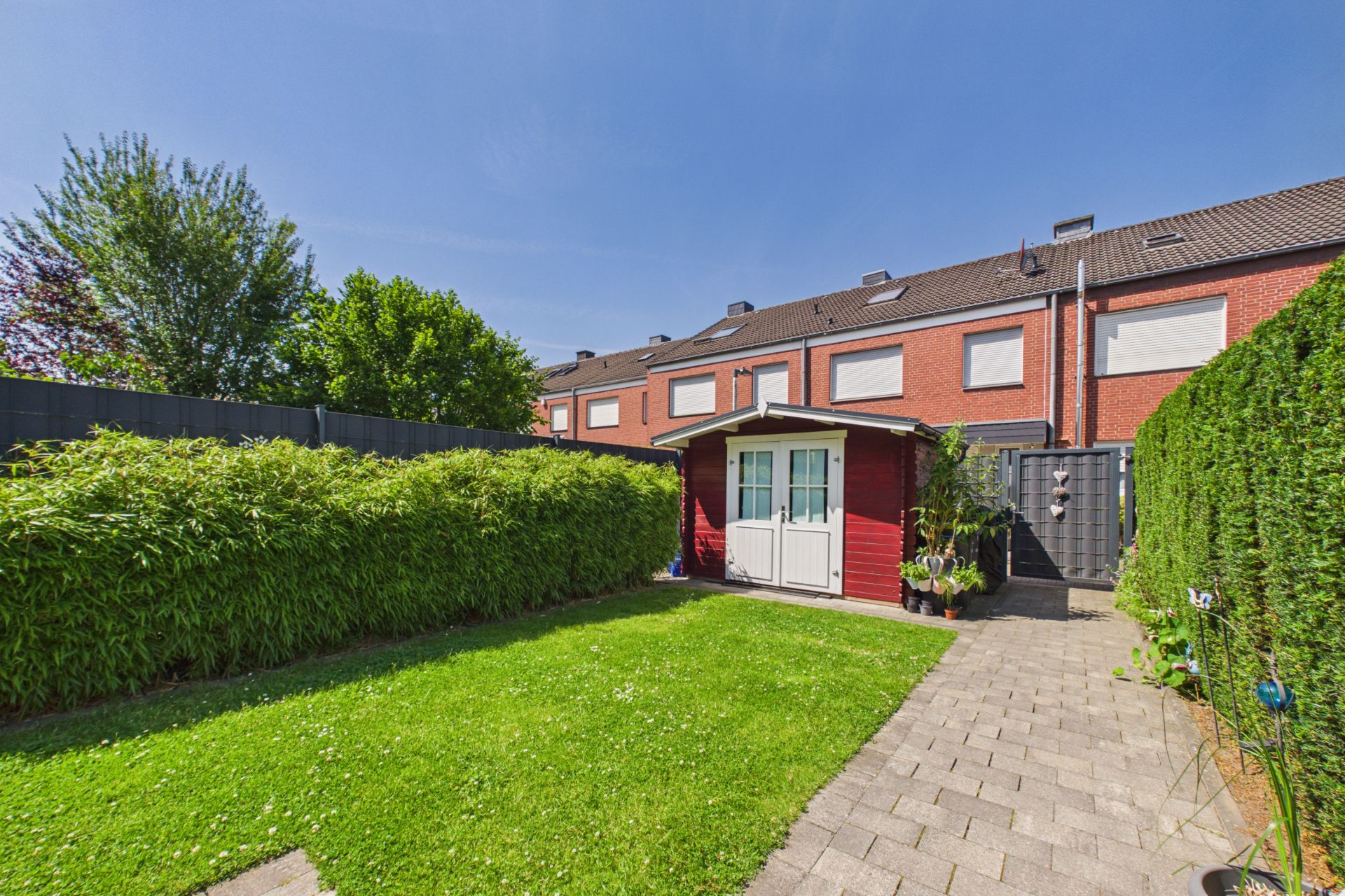 Garten mit Rasen, Weg aus Pflastersteinen und kleinem roten Gartenhaus, umgeben von Hecken.