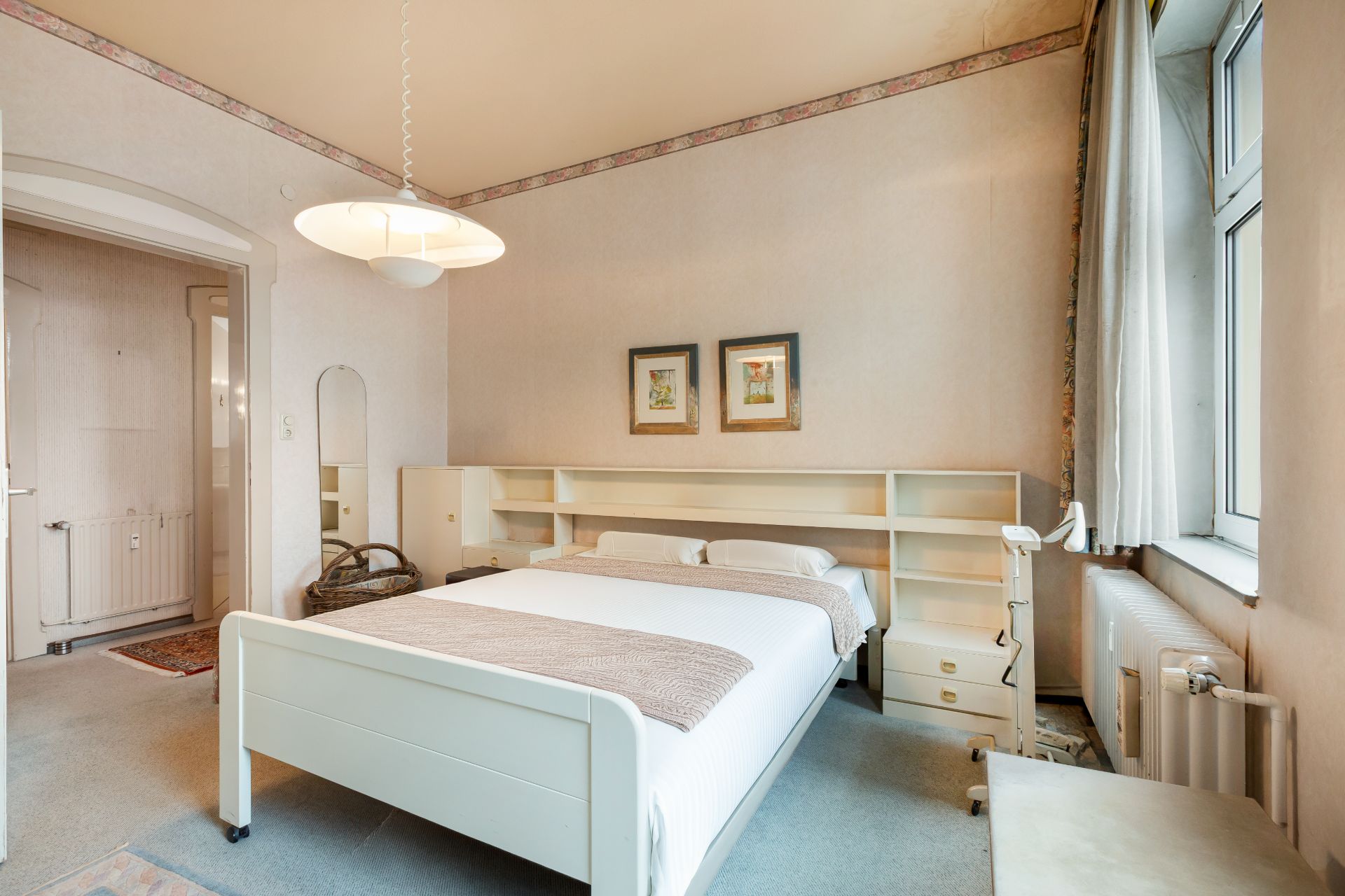 Helles Schlafzimmer mit Doppelbett, Regal hinter dem Bett und großem Fenster auf der rechten Seite.