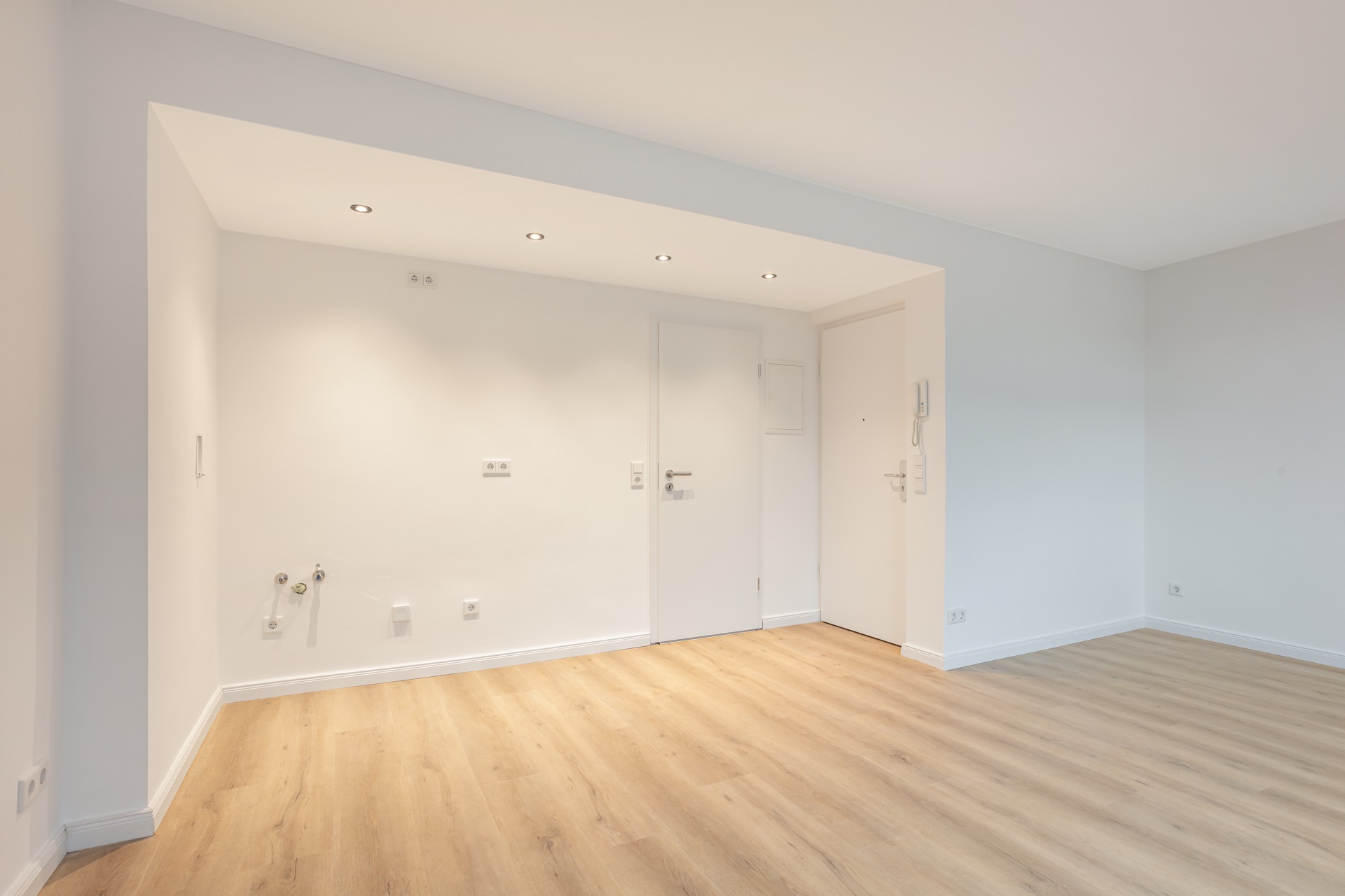 Heller, leerer Raum mit Holzfußboden und weißen Wänden, Türen und Einbauspots in der Decke.