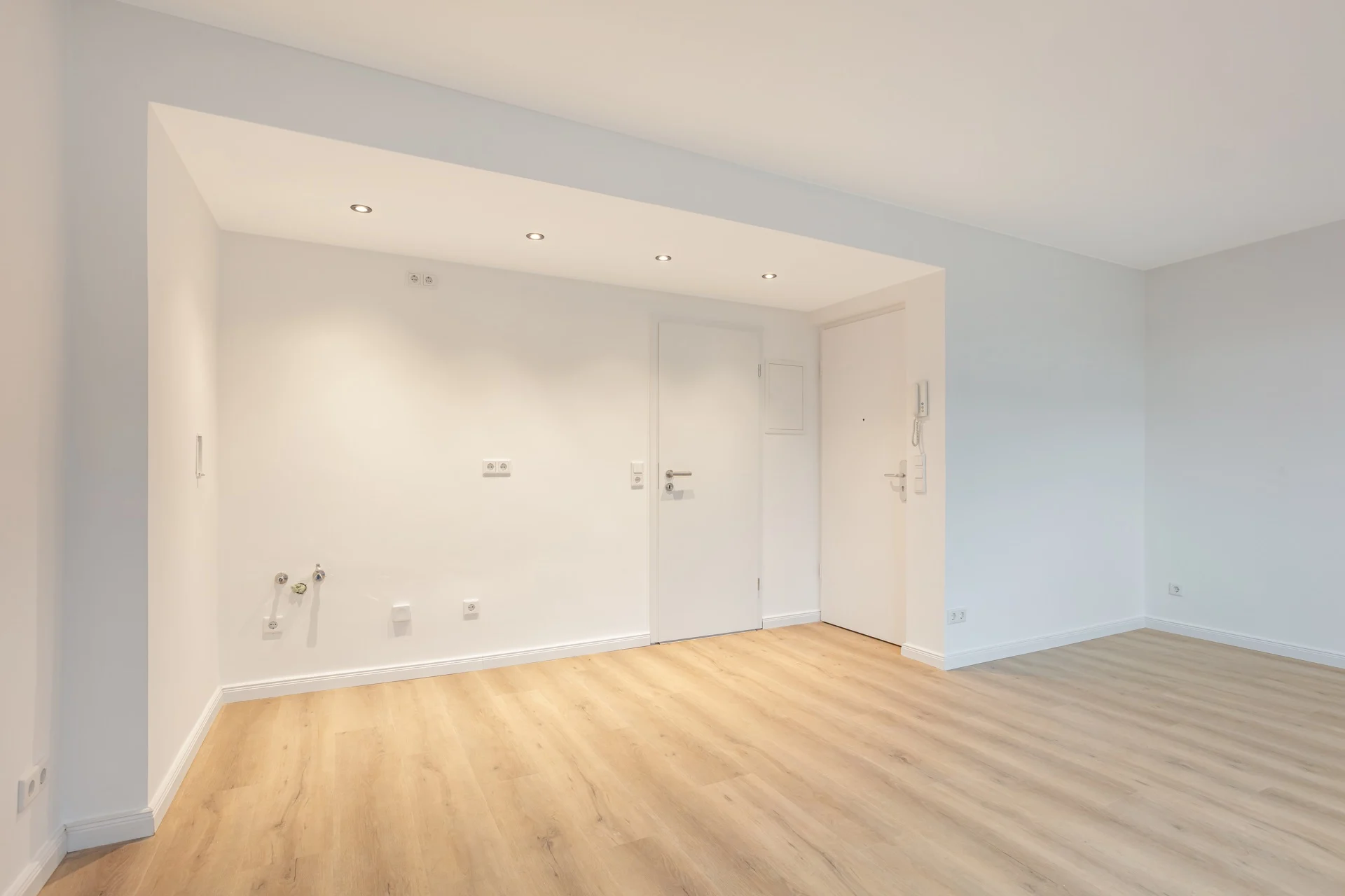 Heller, leerer Raum mit Holzfußboden und weißen Wänden, Türen und Einbauspots in der Decke.