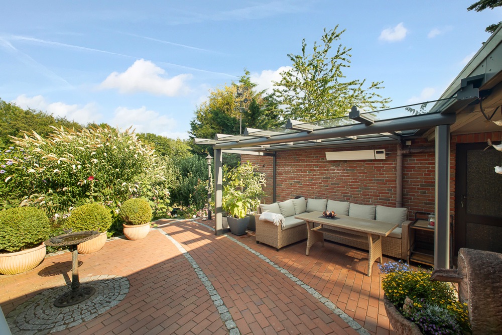 Überdachte Terrasse mit Sofa, Tisch und Pflanzen, angrenzend an einen Garten mit Büschen.
