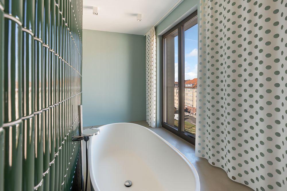 Helles Bad mit freistehender Badewanne, grün gefliester Wand und großem Fenster mit gepunkteten Vorhängen.