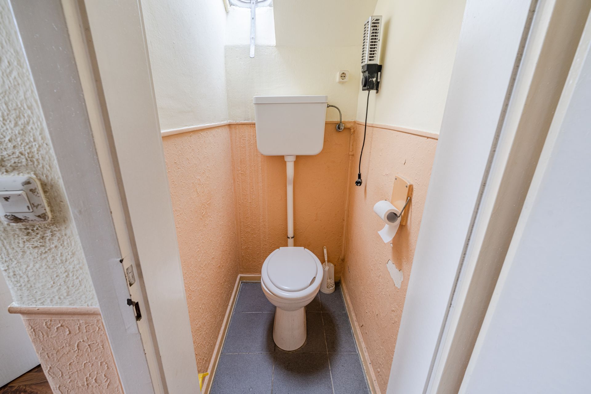 Kleines WC mit Toilette, Wandspülkasten, Toilettenpapierhalter und kleinem Fenster.
