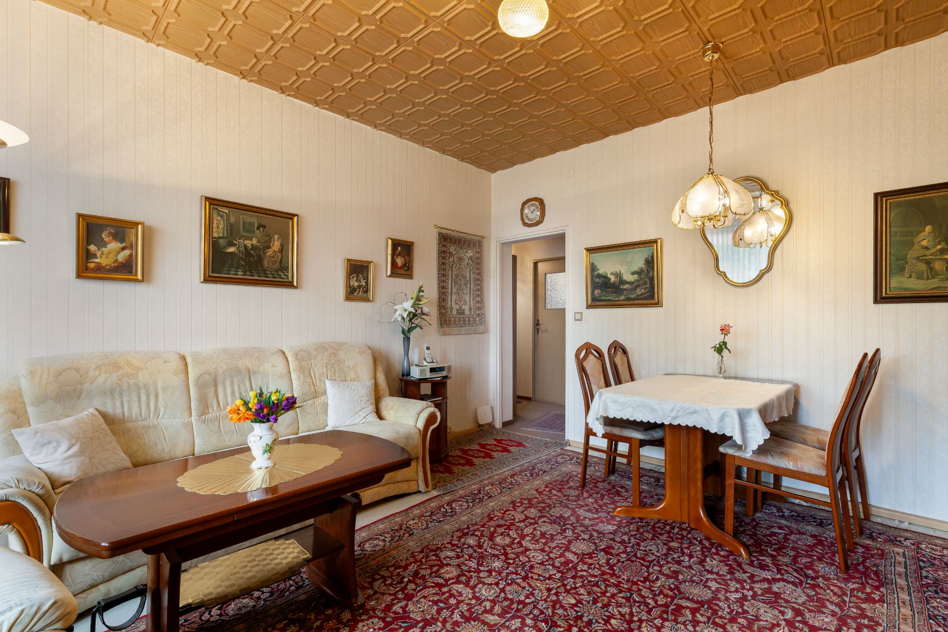 Wohnzimmer mit Teppich, Sofa, Esstisch, Stühlen, Bildern an den Wänden und Deckenlampe.
