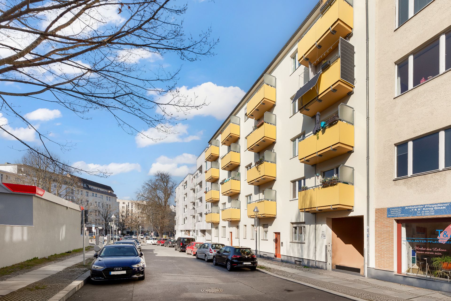 Wohnhaus mit gelben Balkonen an einer ruhigen Straße, mehrere geparkte Autos am Straßenrand.
